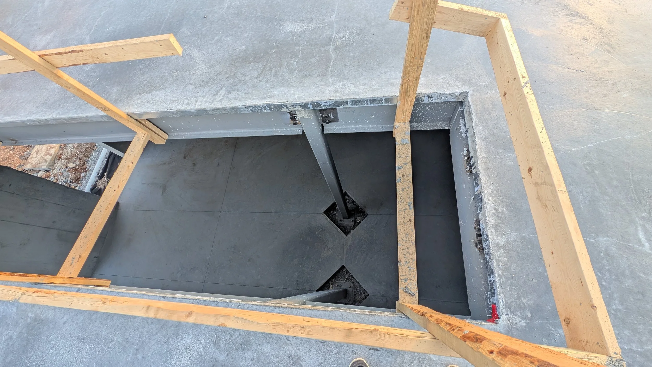 Construction site with a rectangular hole in the concrete floor, supported by wooden framing and metal braces, with black piping inside the hole.