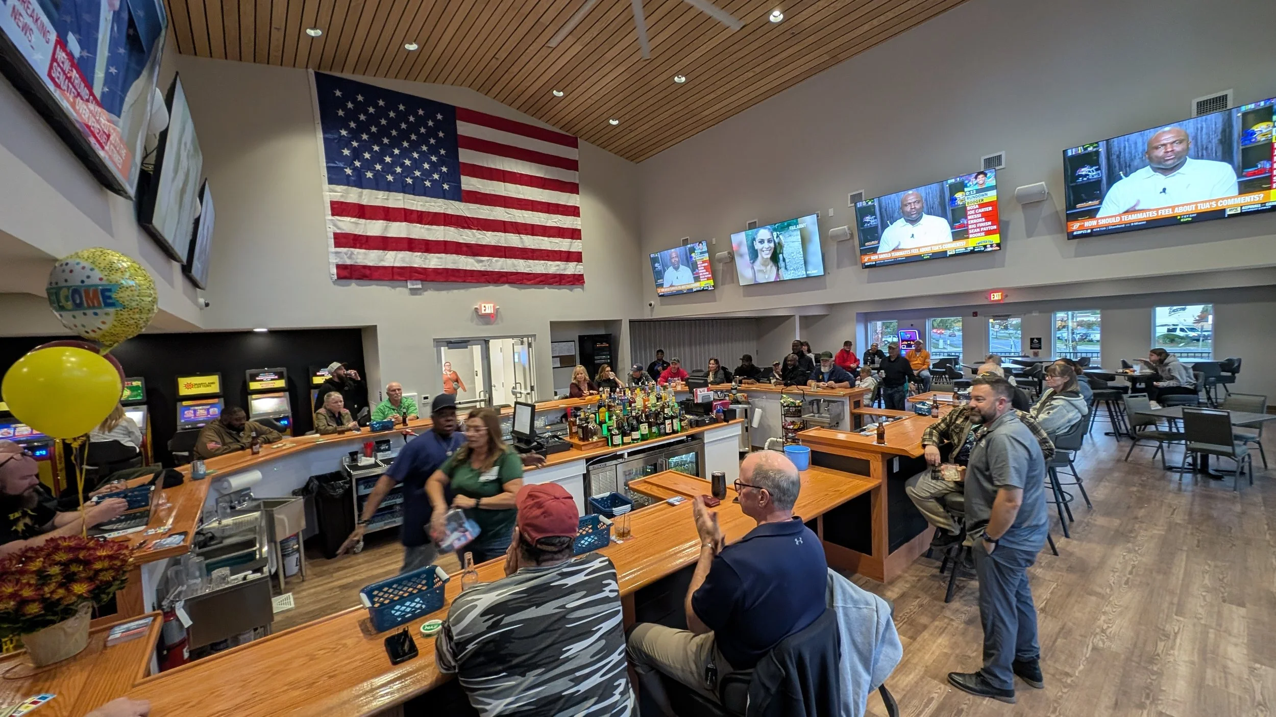 People gathered in a bar or restaurant with a large American flag hanging on the wall and multiple TVs displaying sports and news.
