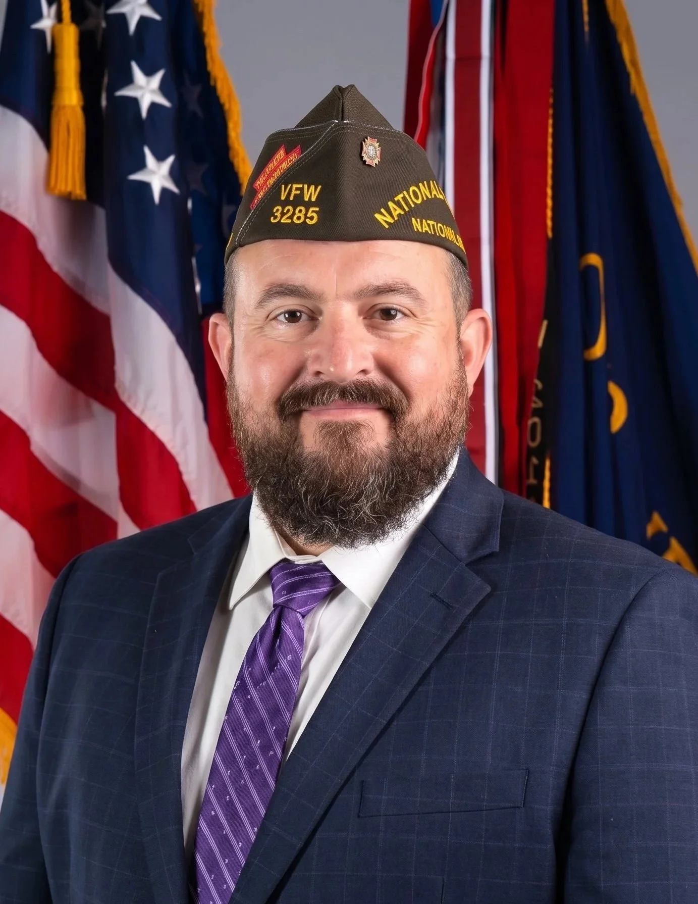 A man with a beard wearing a military-style hat, suit, and a red tie, standing next to an American flag.