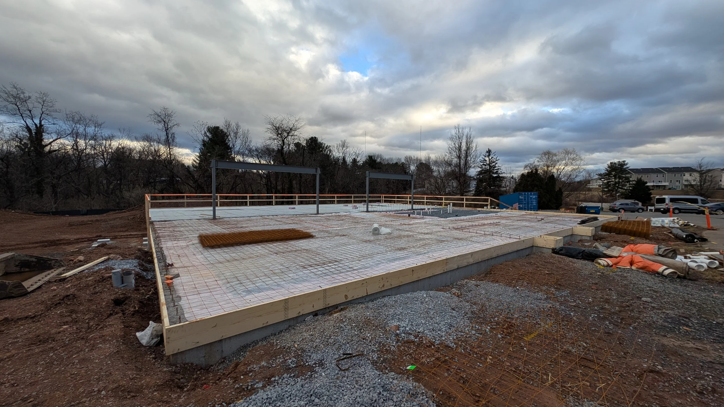 Construction site with concrete slab foundation, surrounded by gravel and dirt, with construction materials and rebar, under a cloudy sky.