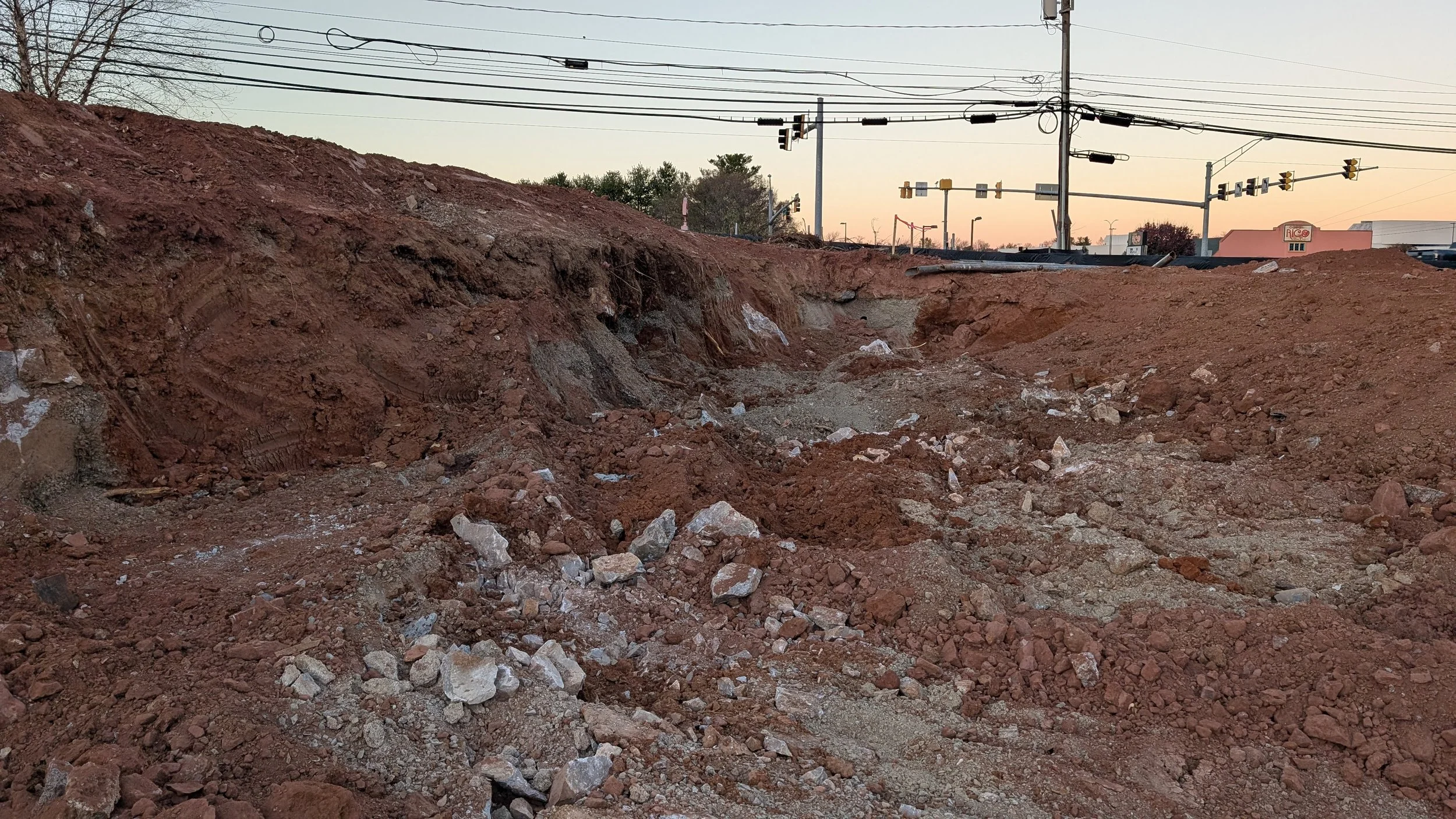Construction site with exposed dirt and rocks, utility lines overhead, traffic signals, and commercial buildings in the background at sunset.