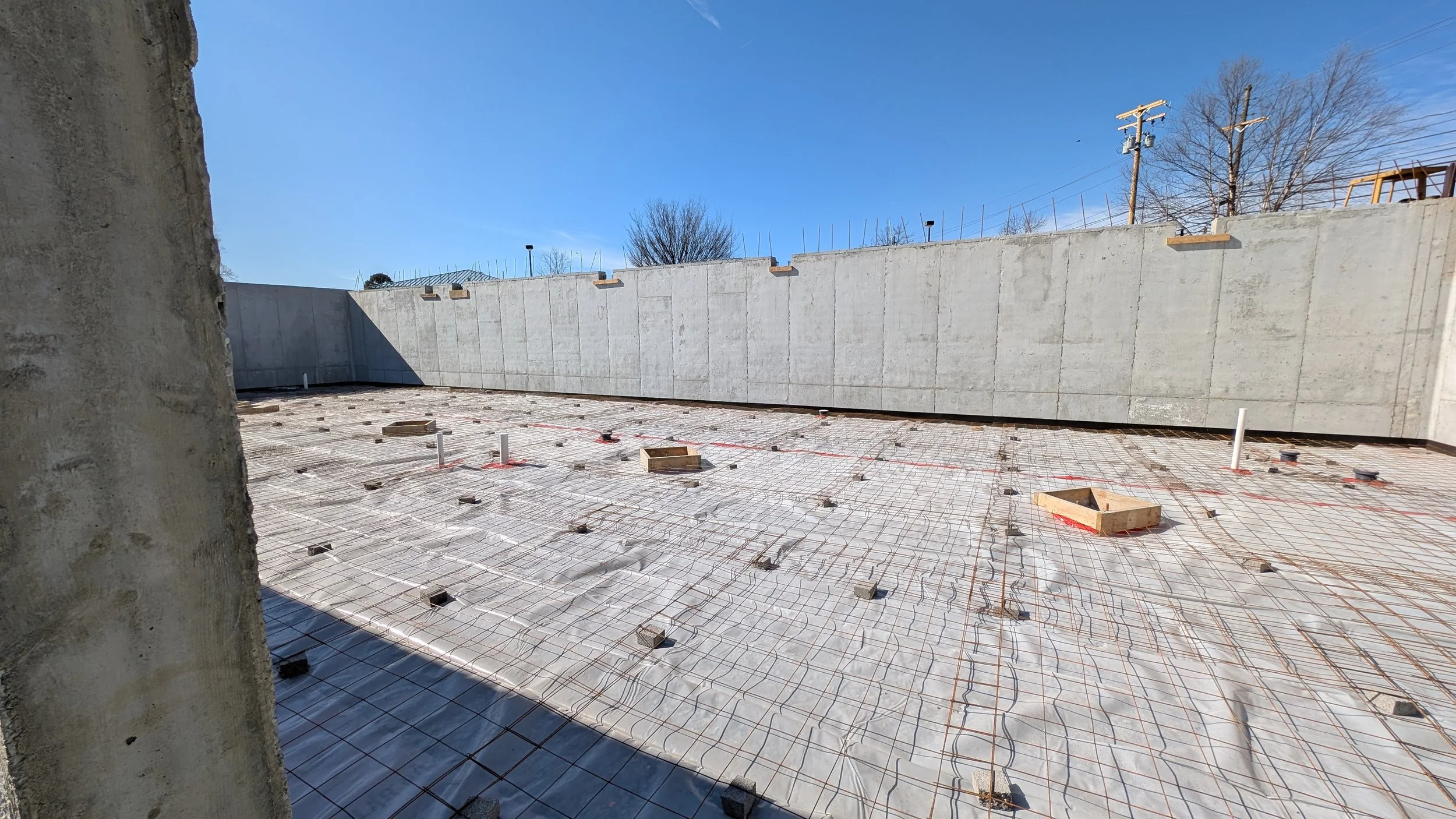 Construction site showing a concrete foundation with rebar grid, pipes, and formwork, enclosed by a tall gray concrete wall with trees and utility poles in the background under a clear blue sky.