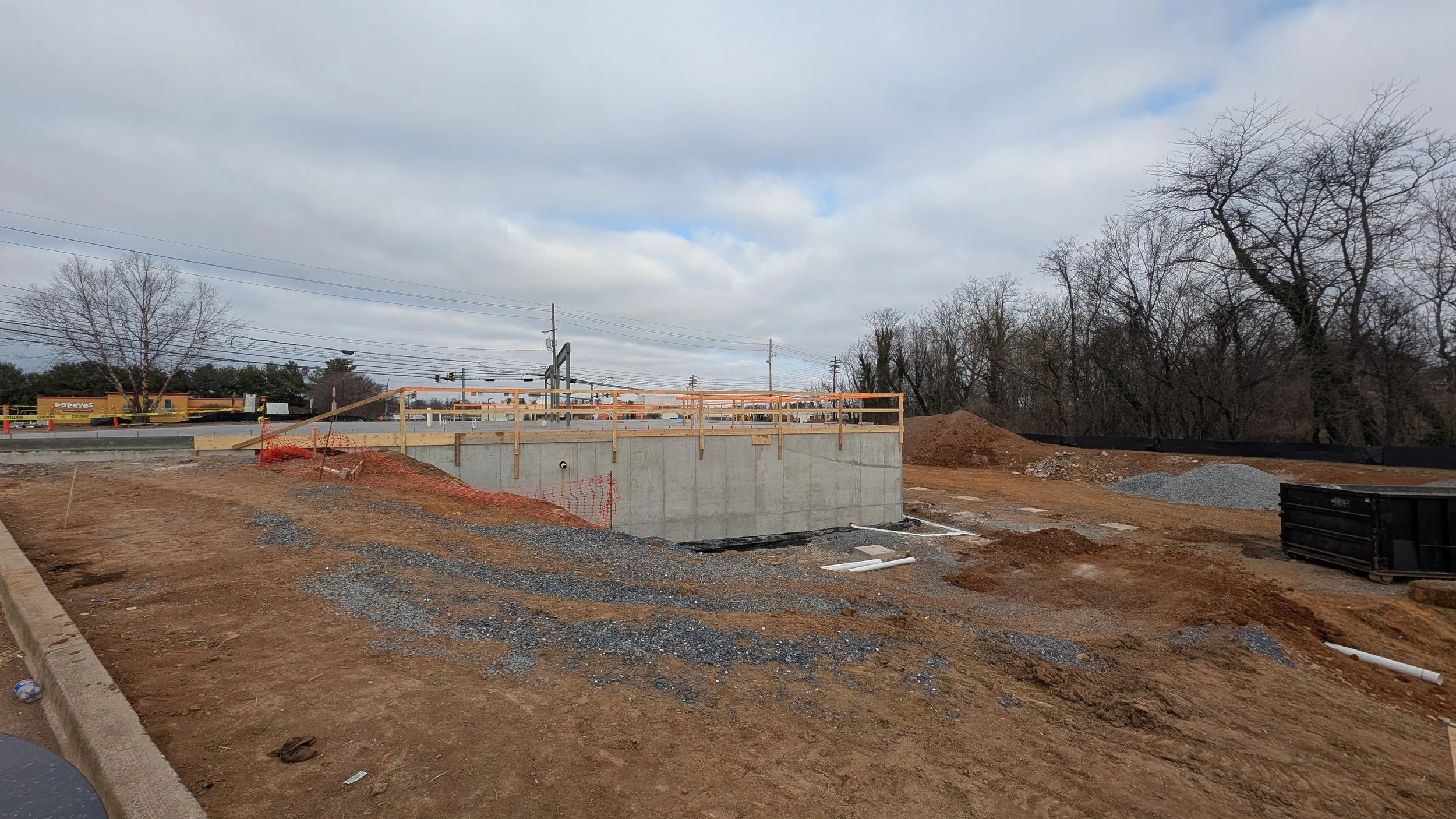 Construction site with a concrete structure, orange safety fencing, and piles of dirt and gravel. There are leafless trees and power lines in the background under a cloudy sky.
