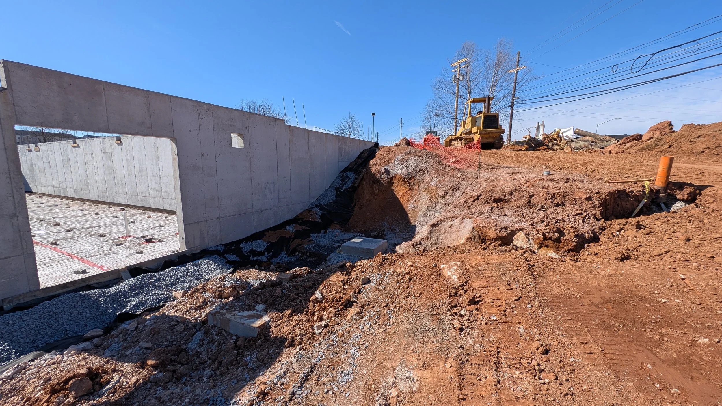 Construction site with a large concrete wall, dirt, and construction equipment, including a small bulldozer, under a clear blue sky.
