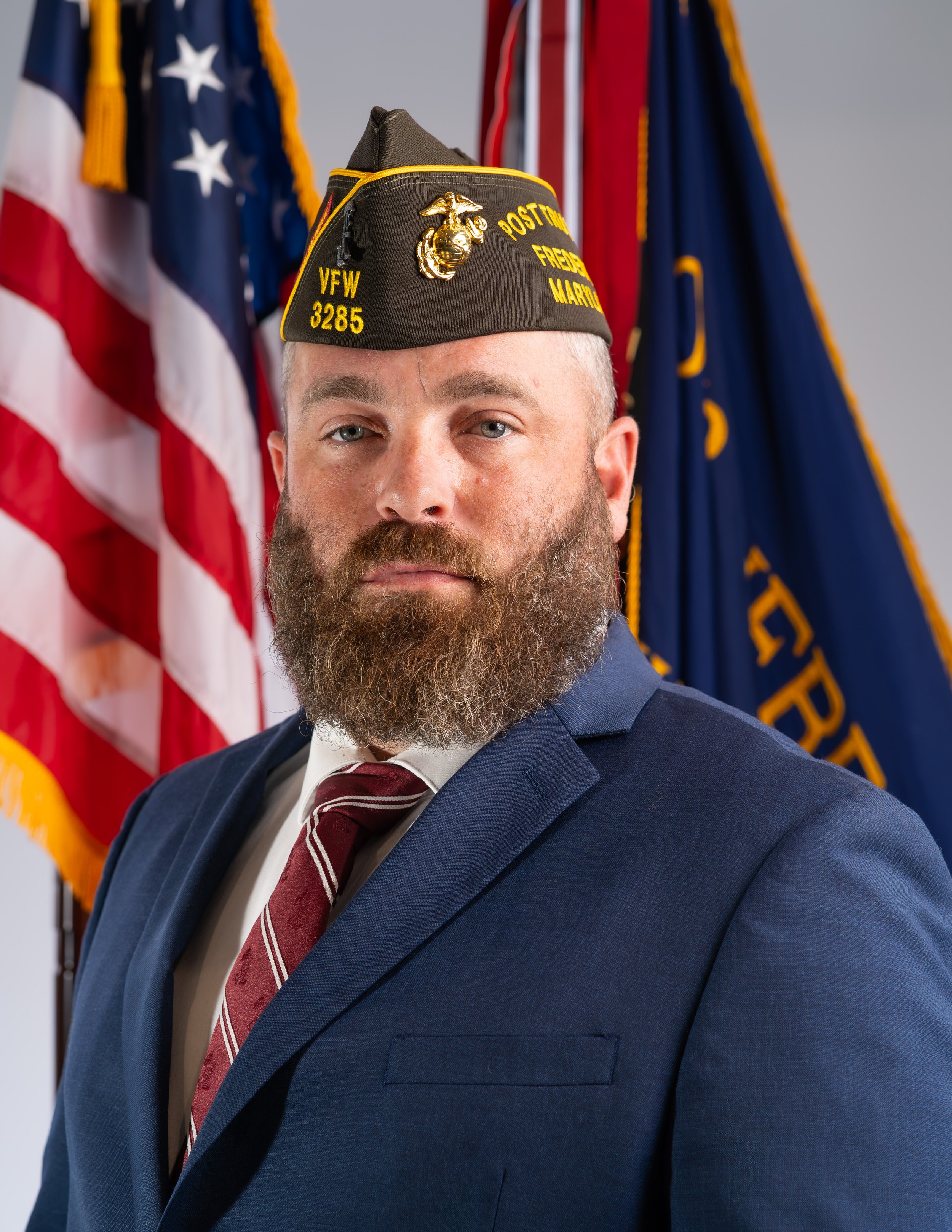 A man with a beard and curly hair smiling, wearing a U.S. Marine Corps vest with various patches and ribbons, a black Veteran of Foreign Wars (VFW) cap, and standing next to an American flag.