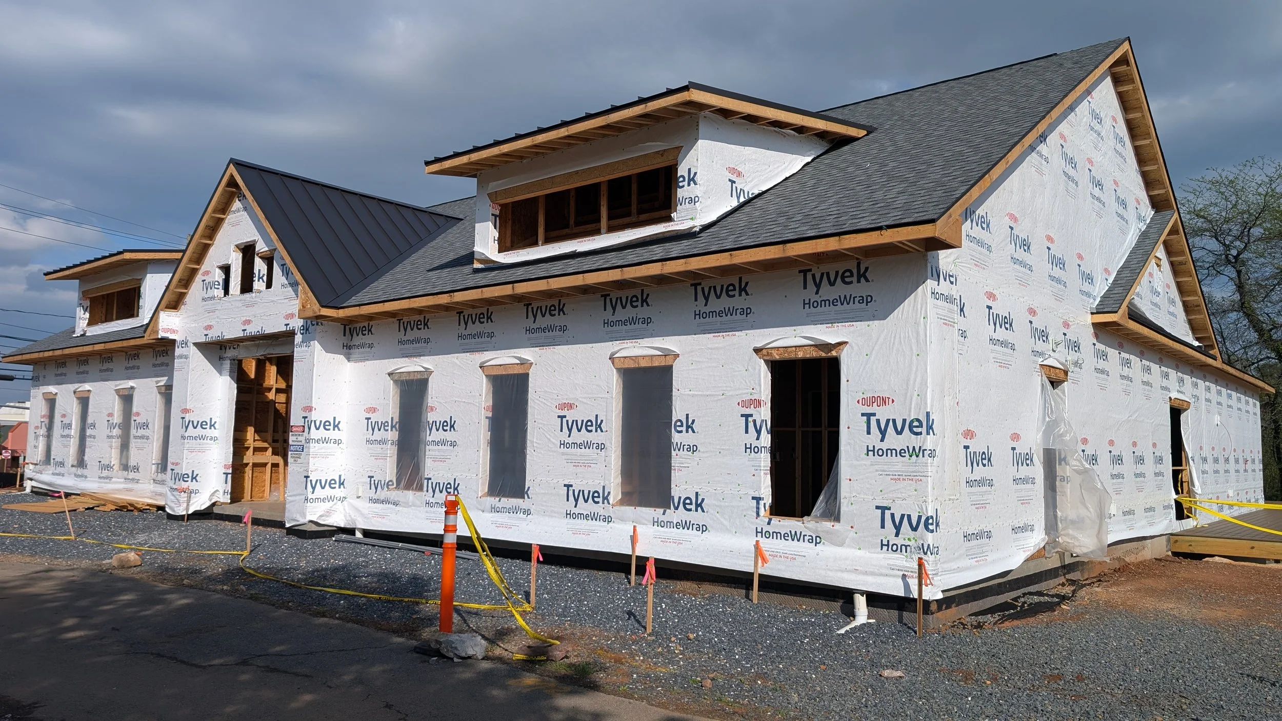 A house under construction covered with Tyvek house wrap, with some windows and opening frames installed, and a dark gray roof partially completed.