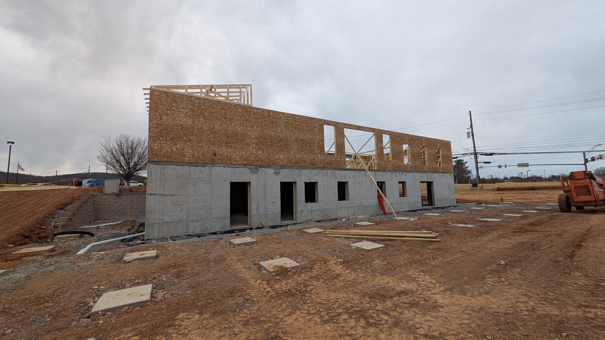 Construction site of a two-story building with a concrete lower level and wooden upper frame, on a dirt lot under a cloudy sky.