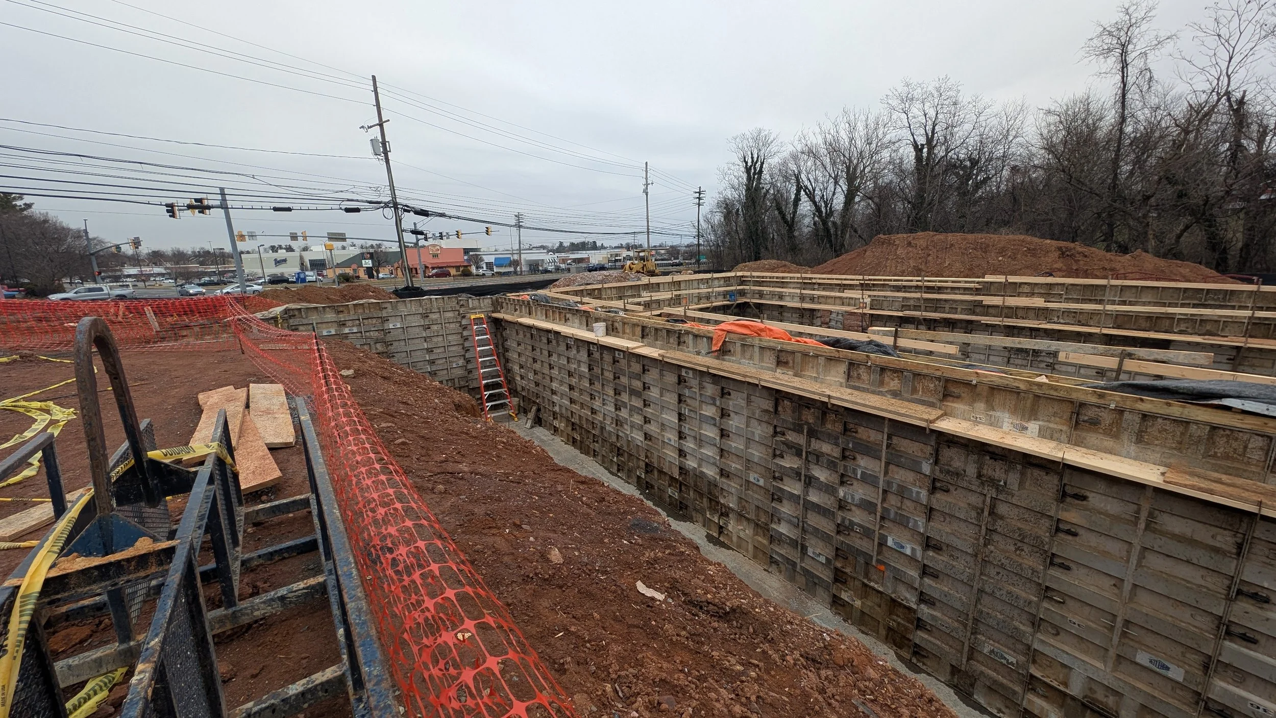A construction site with deep excavation for a building foundation, wooden forms in place, and construction equipment, with a parking lot and stores in the background.