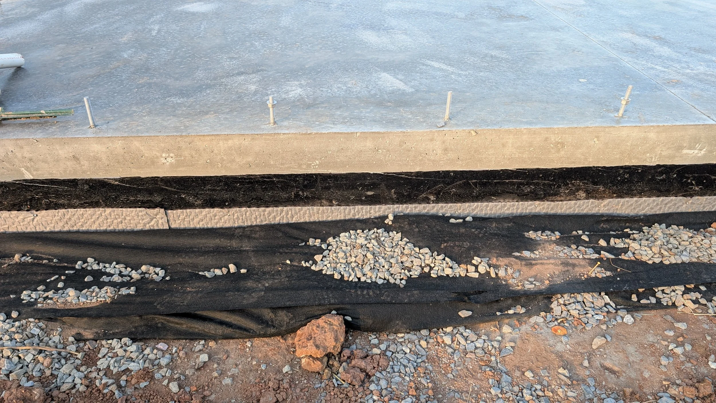 Construction site showing a concrete slab, waterproofing membrane, gravel, and dirt with small rocks and soil.