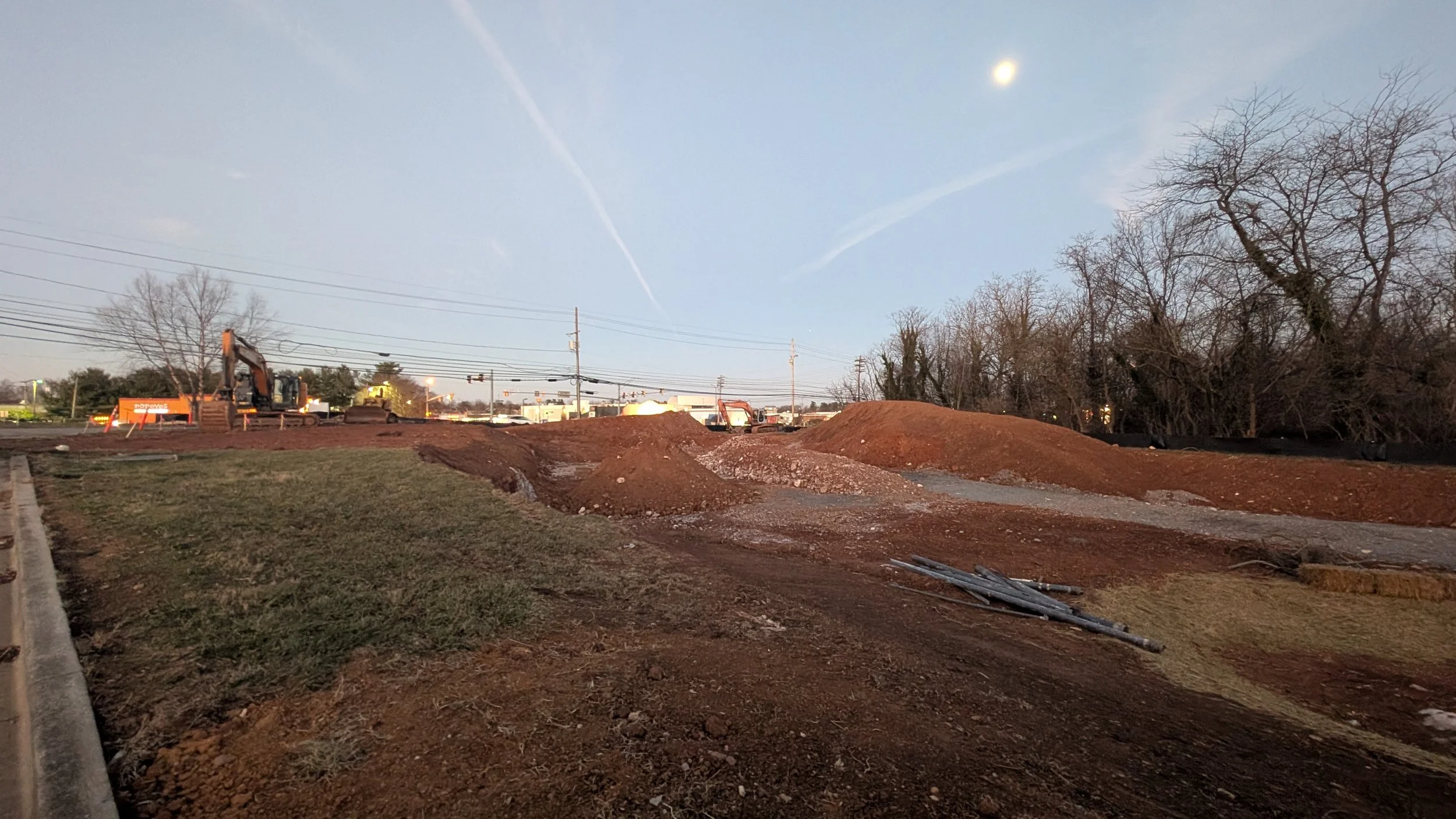 Construction site with dirt mounds, excavator, and construction equipment under a clear sky with visible moon and trees in the background.