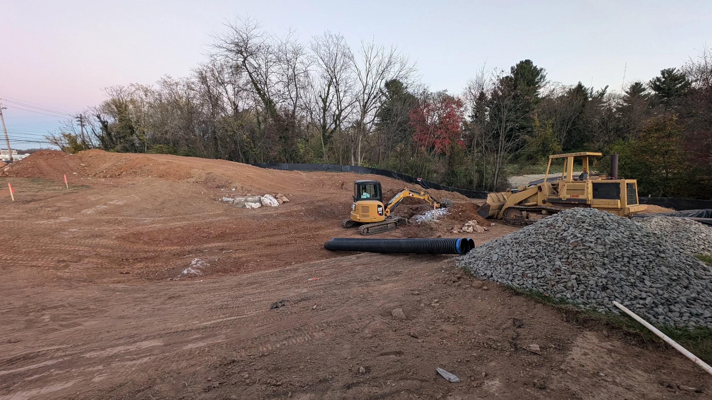 Construction site with two bulldozers, piles of gravel, and a large black pipe on the ground, with trees in the background.