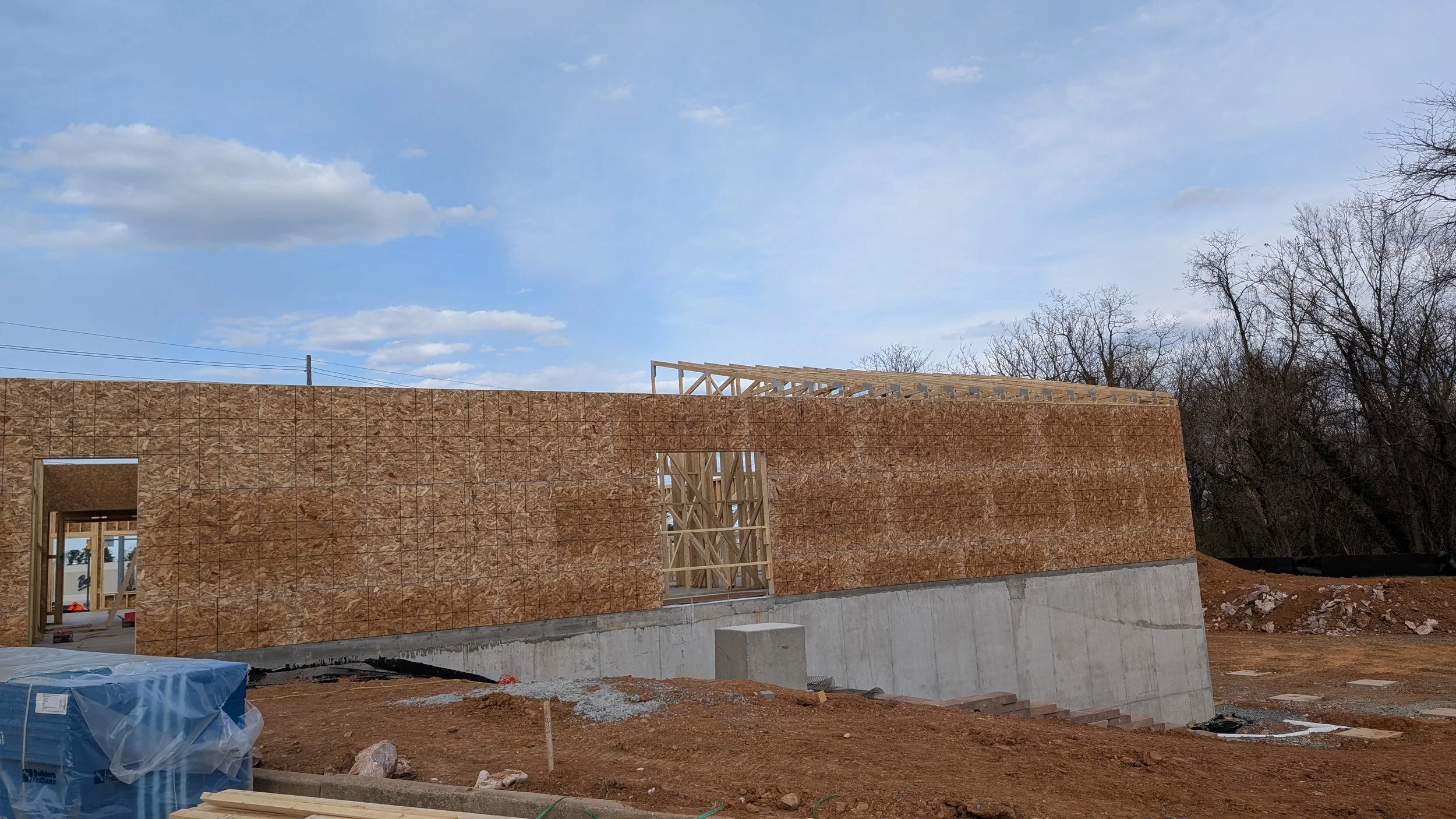 Construction site with wooden framing on top of a concrete foundation, with a partially built exterior wall made of oriented strand board (OSB) sheathing, under a partly cloudy sky.