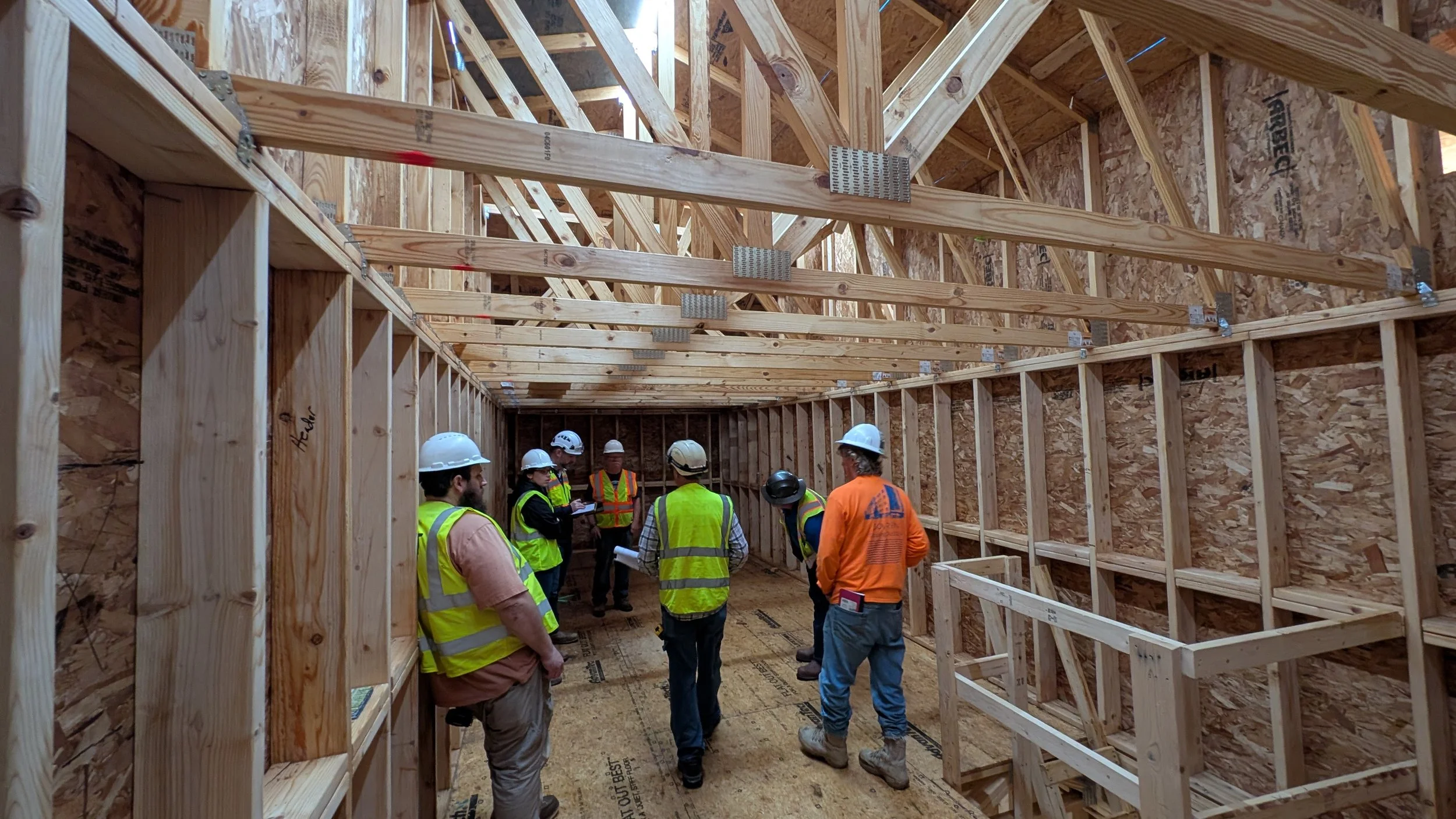 Construction workers in helmets and safety vests inside a wooden framing of a building under construction.