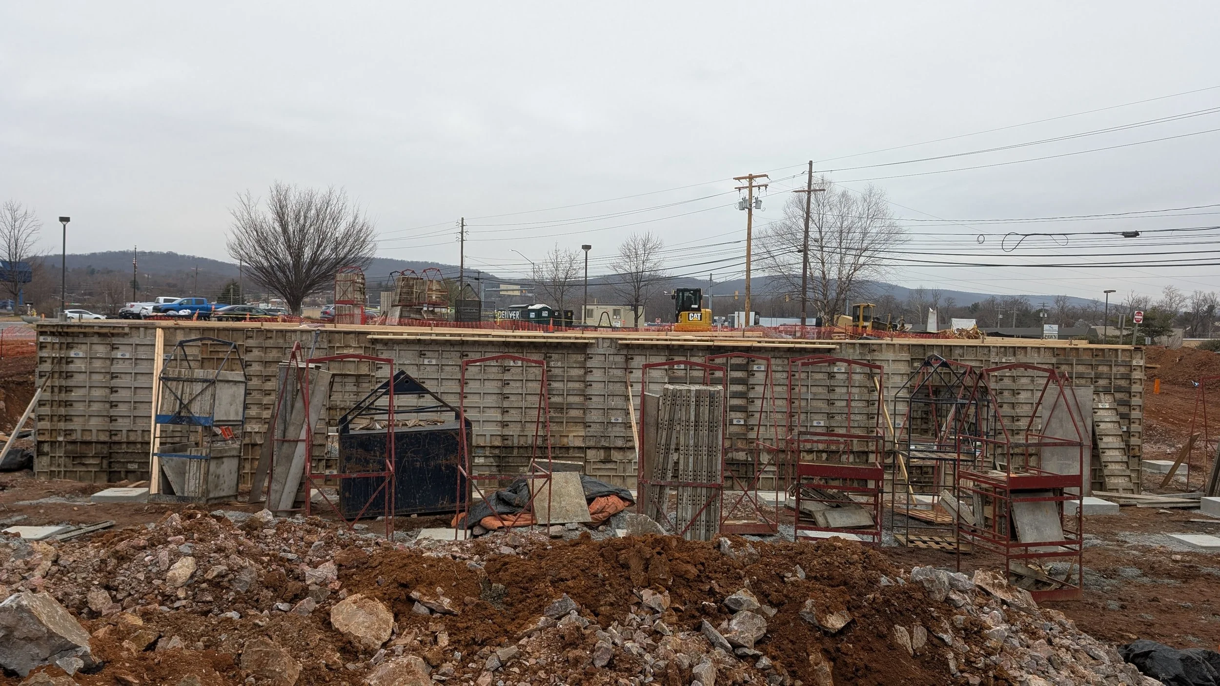 Construction site with concrete forms, scaffolding, and construction materials in the foreground. A retaining wall is built, with construction equipment and vehicles in the background against a cloudy sky.