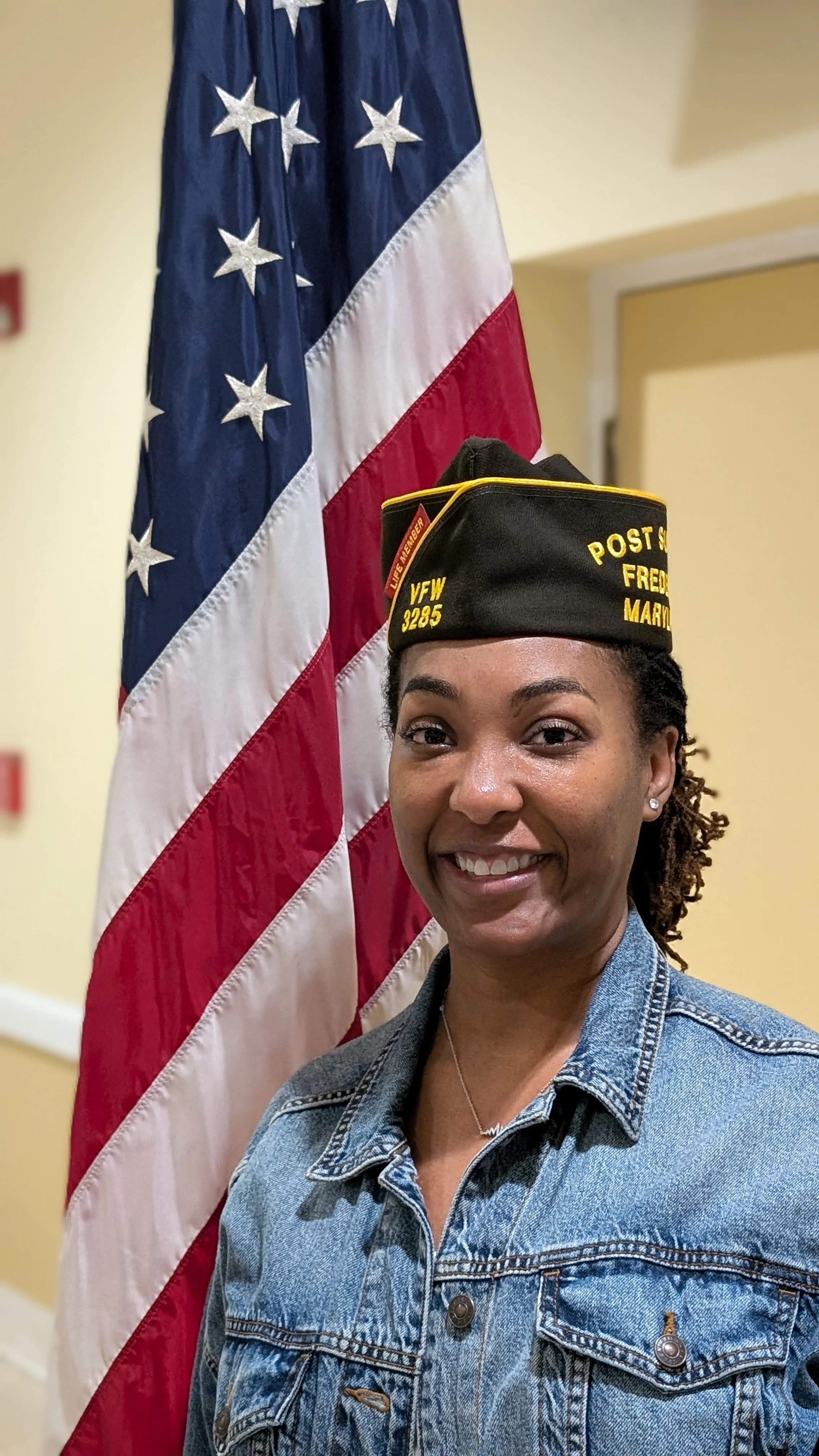 A woman in a uniform with a cap that says 'Post Service FRED MARSHA' standing next to an American flag.