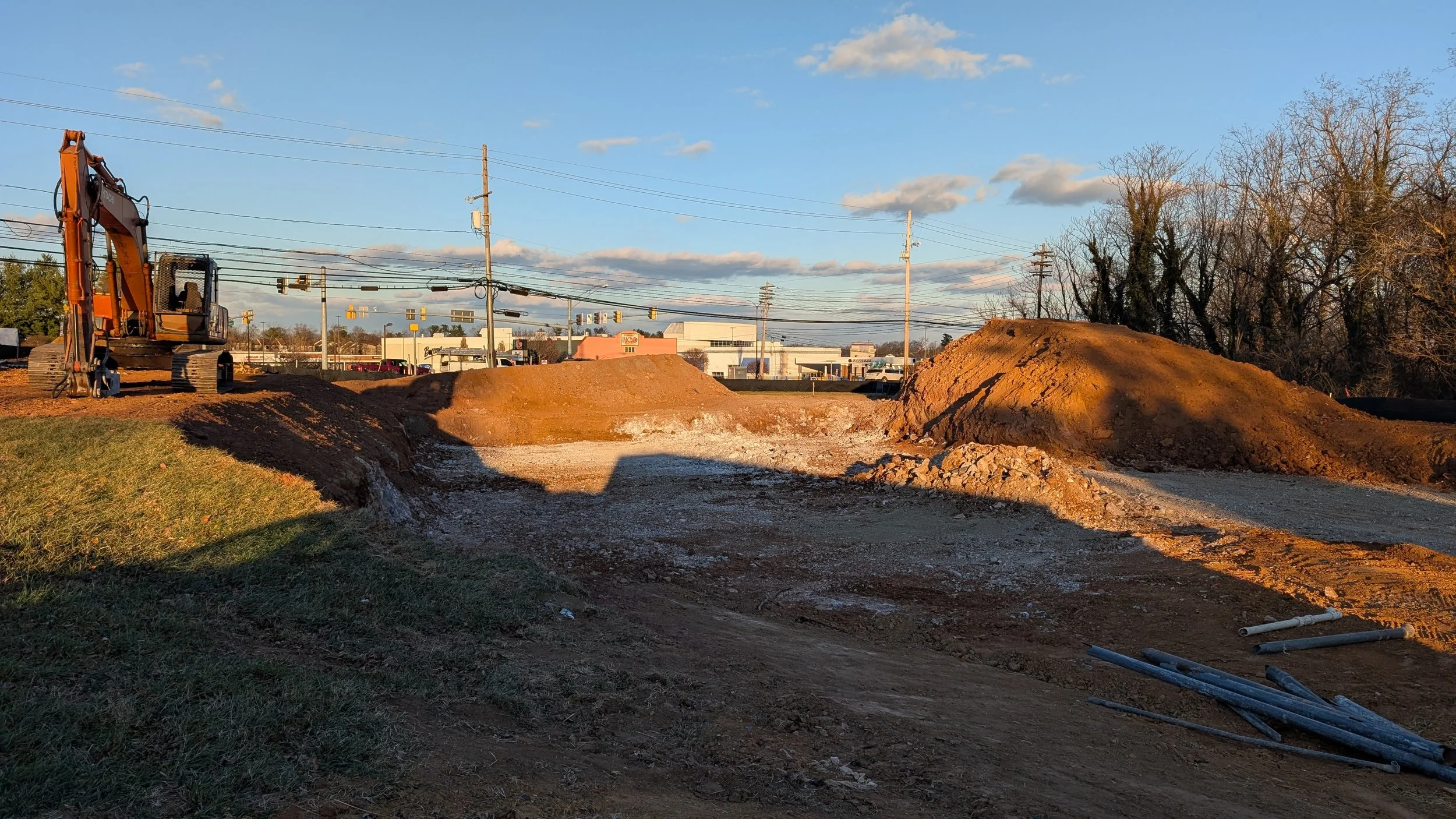 Construction site with a large excavator on the left and dirt piles in the center and right, with a background of commercial buildings, power lines, trees, and a partly cloudy sky.