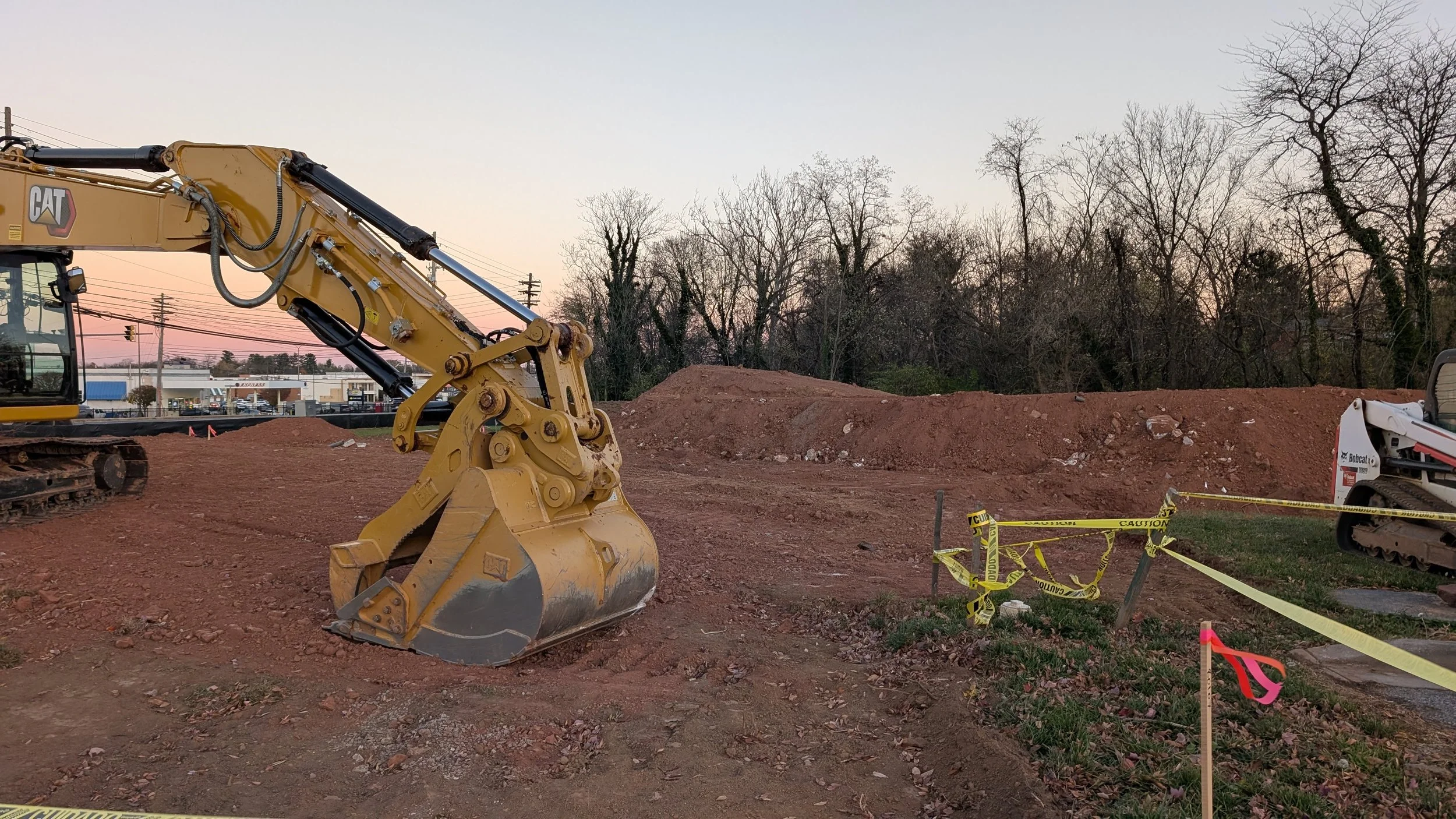 Construction site with excavator and piles of dirt, surrounded by yellow caution tape and trees in the background during sunset.