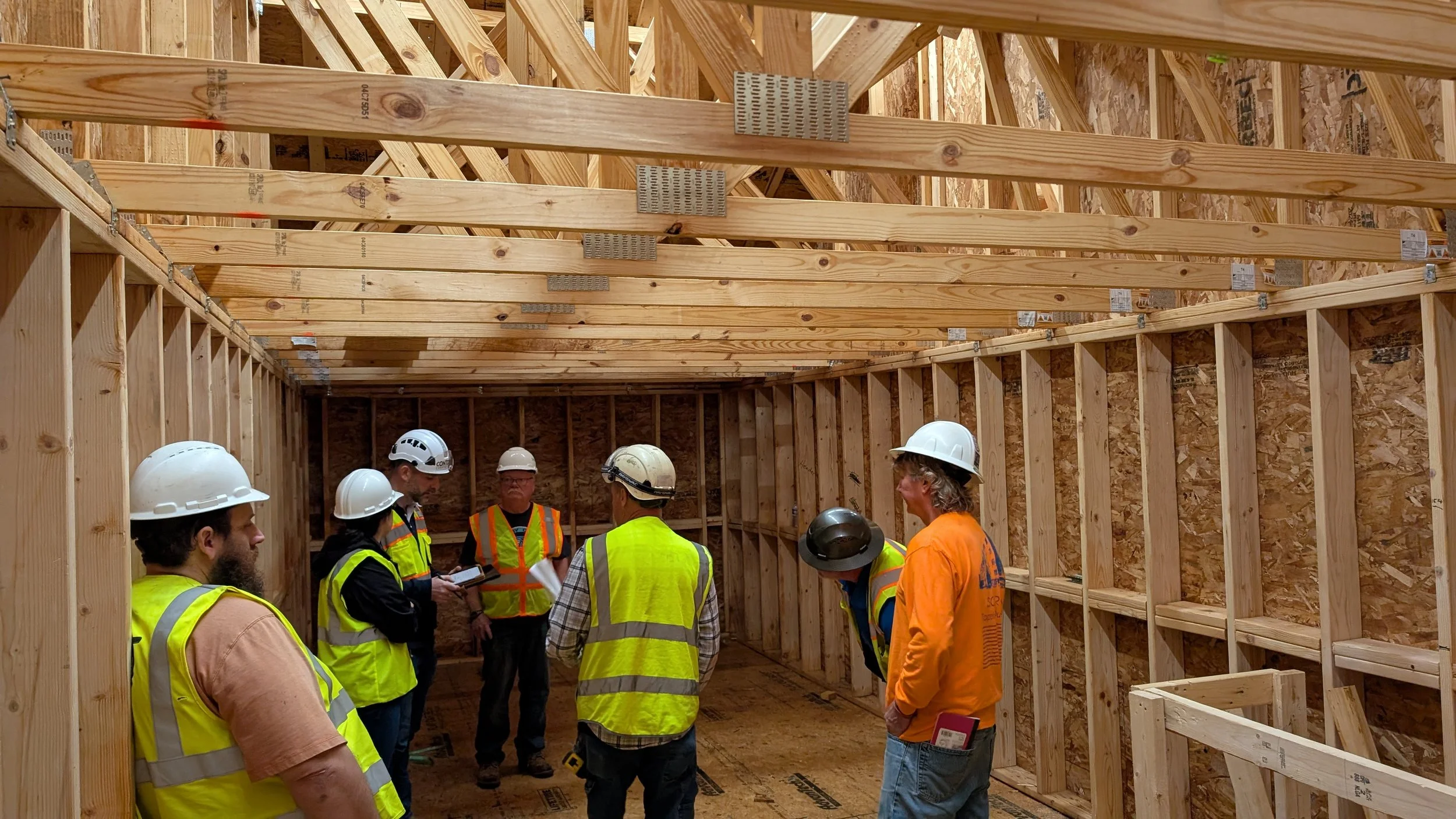 Construction workers and supervisors wearing safety helmets and vests inside a partially built wooden structure, discussing, with wooden framing and beams visible.
