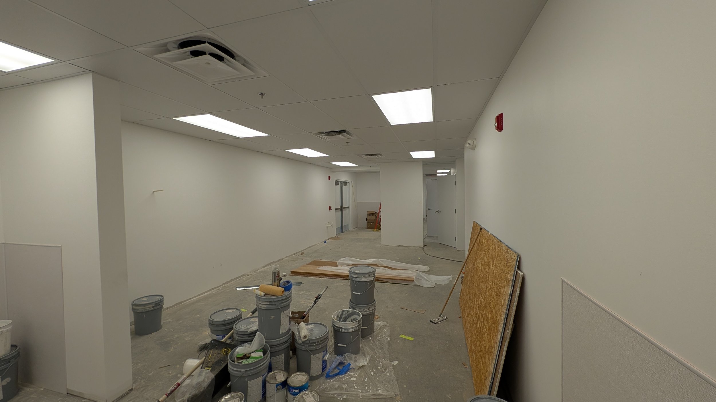 Interior of a room under renovation with construction supplies, paint buckets, and tools on the floor, white walls, and ceiling with fluorescent lights.