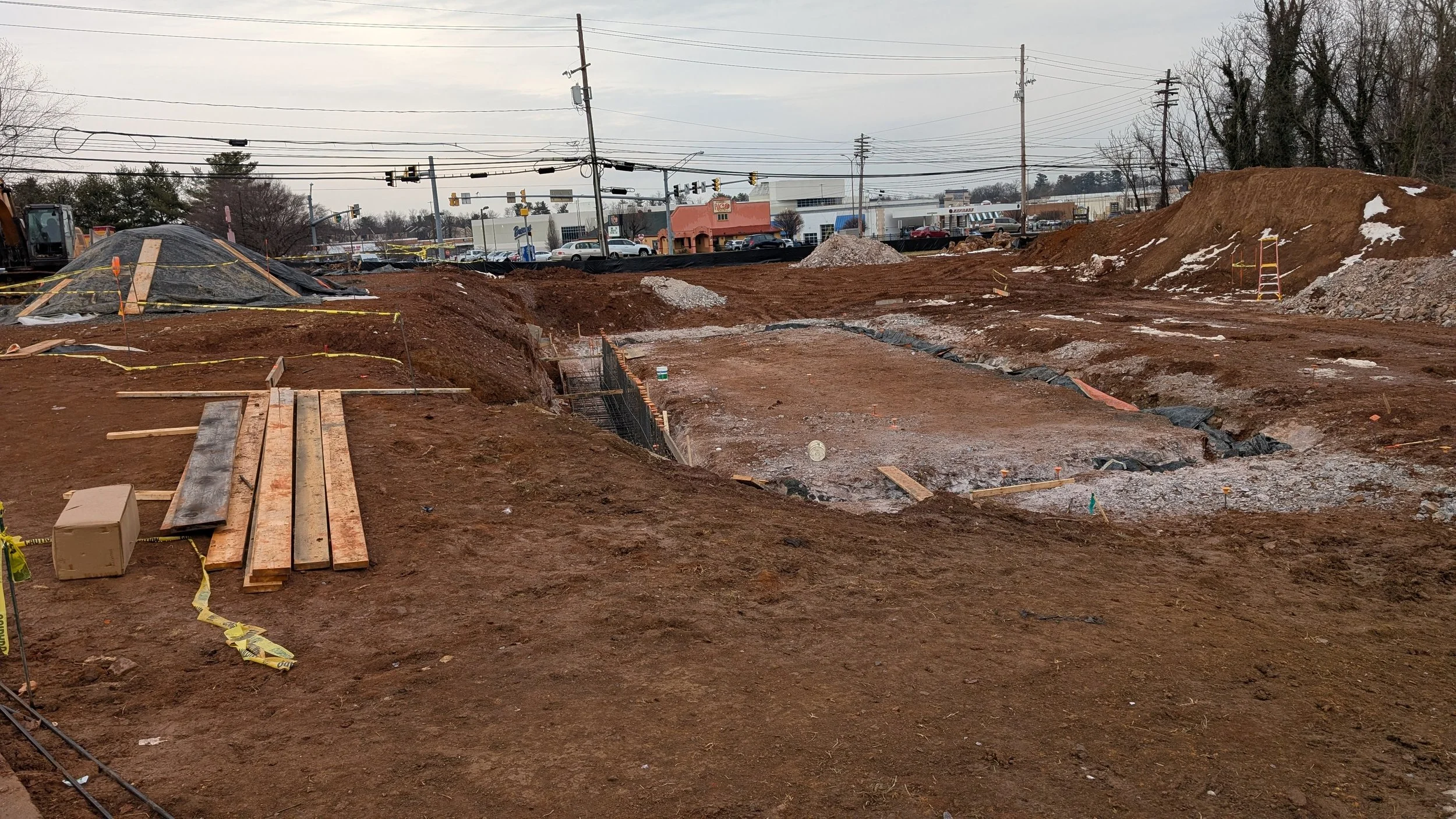A construction site with exposed dirt, trenches, wooden planks, and construction materials. In the background, there are buildings, power lines, and traffic lights.