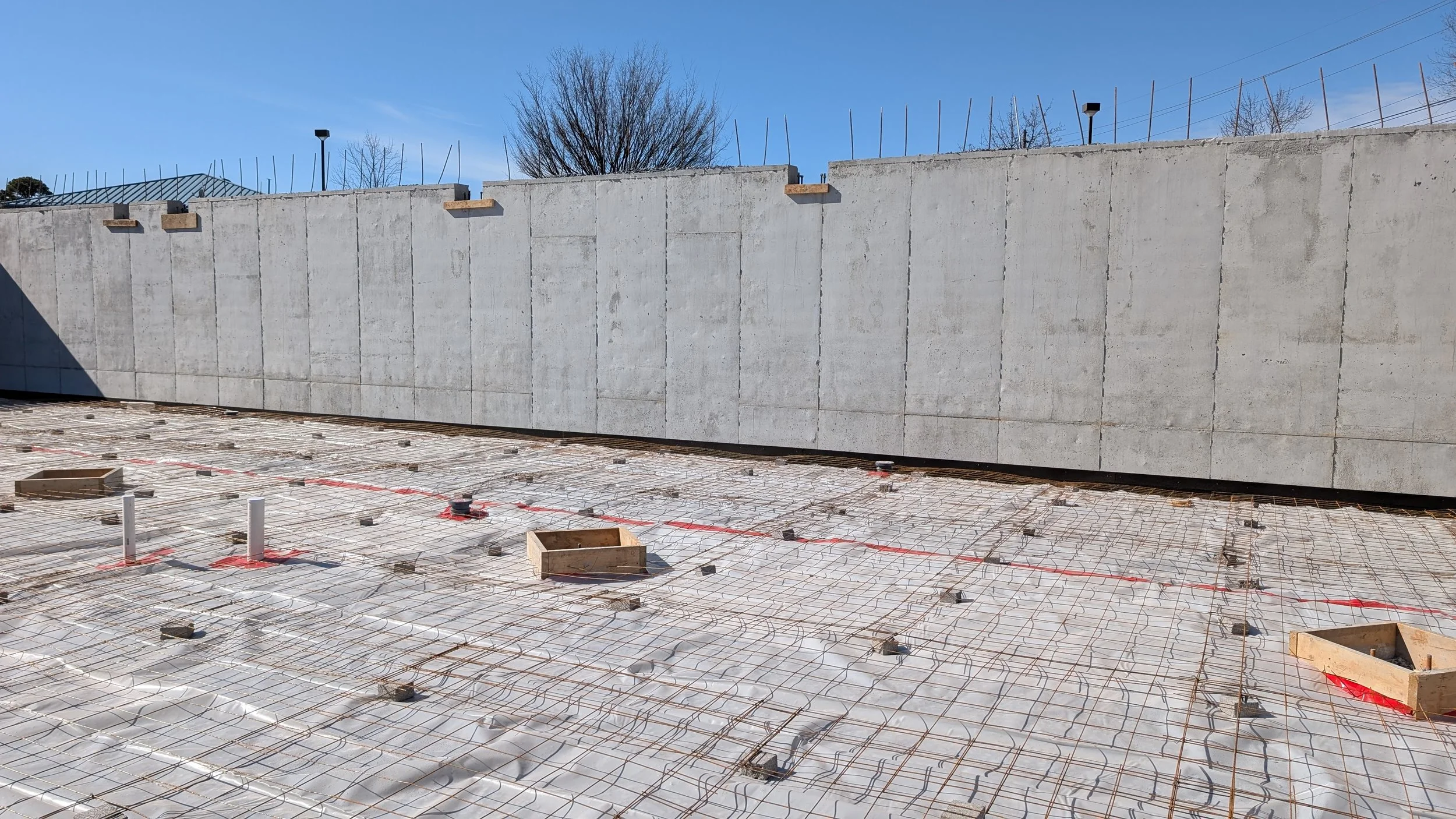 Construction site with concrete wall and rebar grid on ground for concrete pouring, with some wooden boxes and PVC pipes sticking out.
