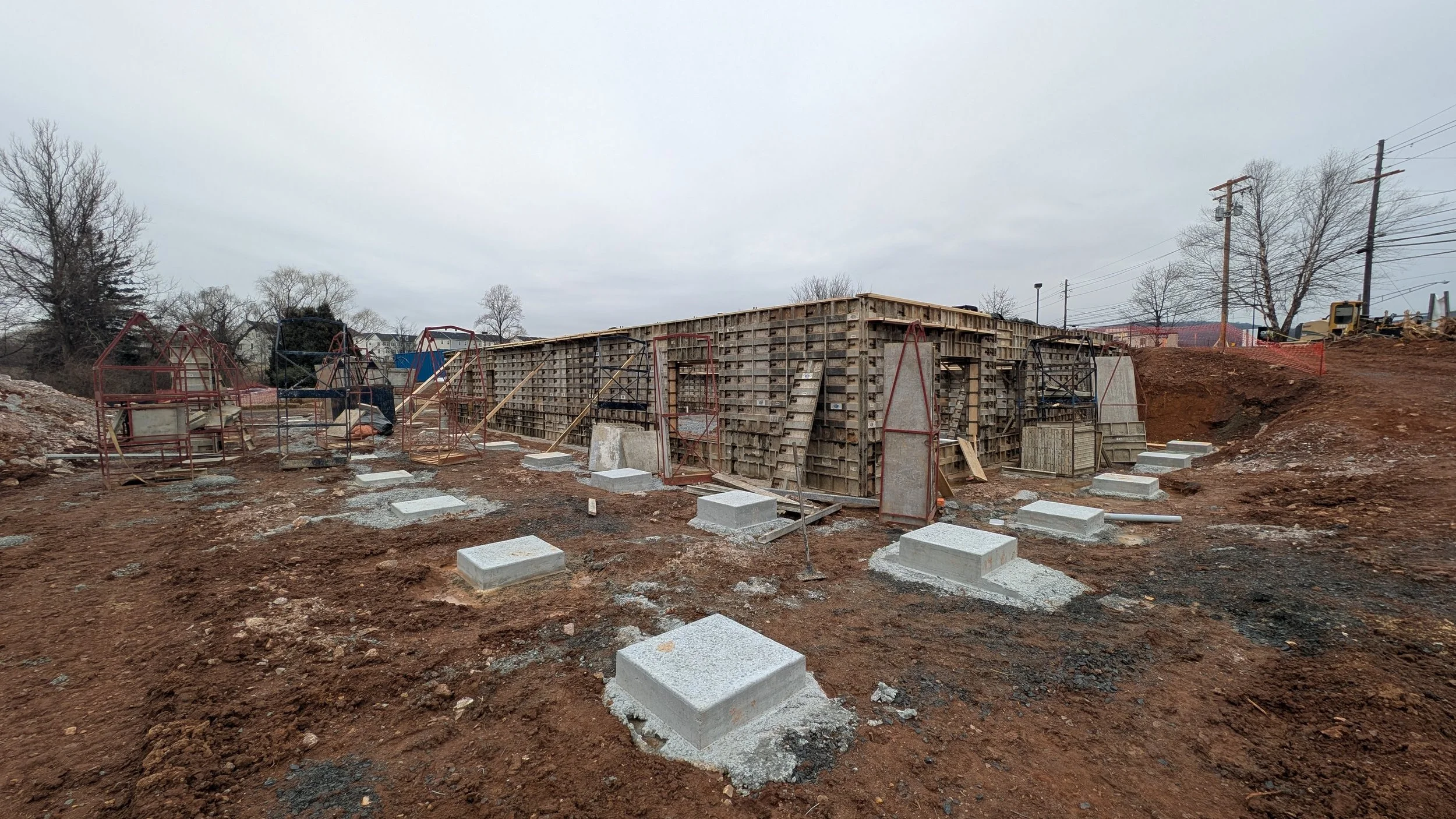 Construction site with partially built concrete structure, rebar, and foundation blocks on muddy ground, with trees and utility poles in the background.