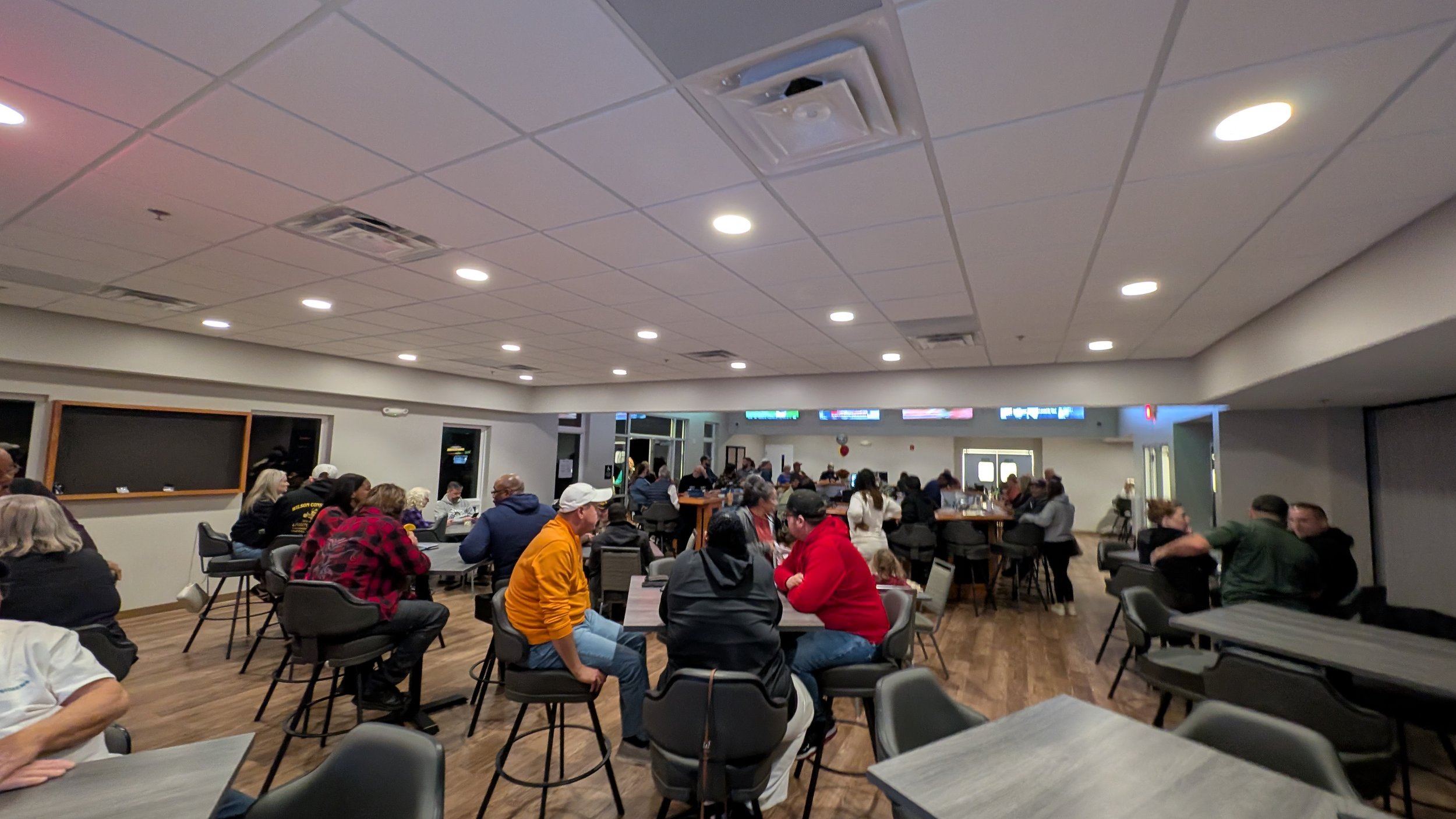 People sitting and socializing in a large, well-lit indoor space with tables and chairs, possibly a community center or event hall.