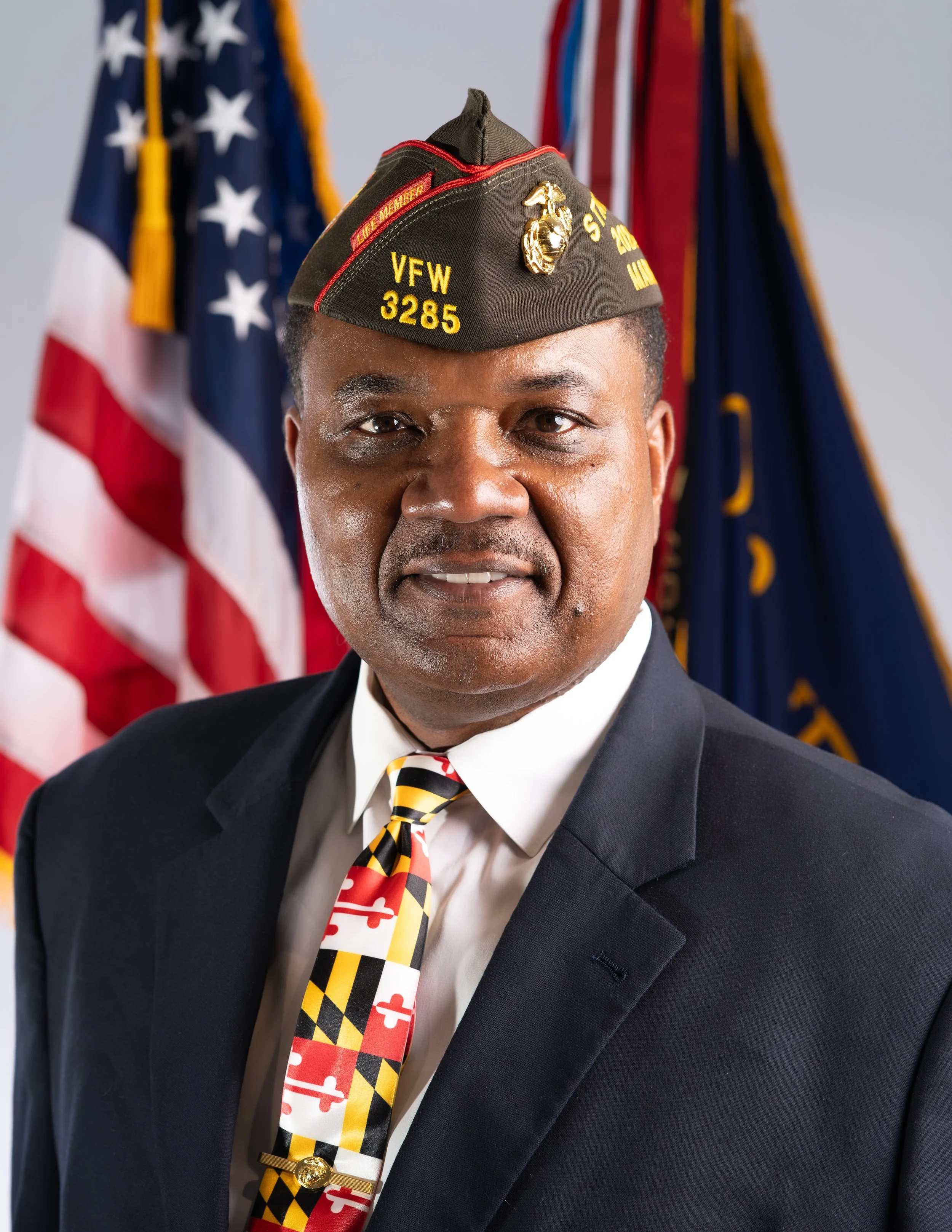 A man in a navy suit and white shirt, wearing a black military-style hat with yellow embroidery reading 'Life Member VFW 3285' and a Marine Corps emblem. He has a patriotic tie with Maryland flag colors and a pin resembling a flag on his lapel. An American flag is partially visible on the right side of the image.
