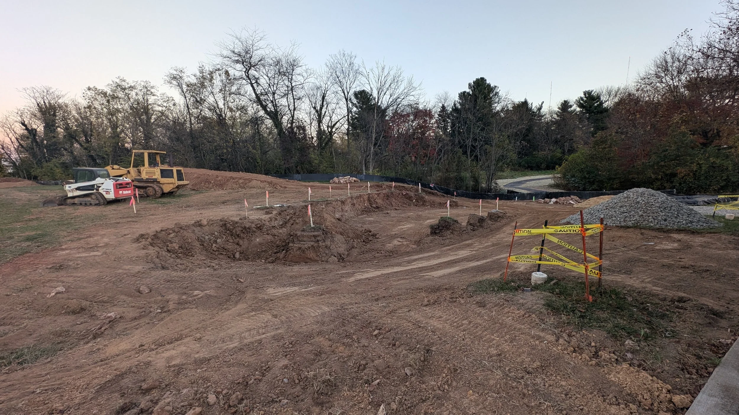 Construction site with bulldozer and excavator on dirt, with caution tape and piles of gravel, surrounded by trees.