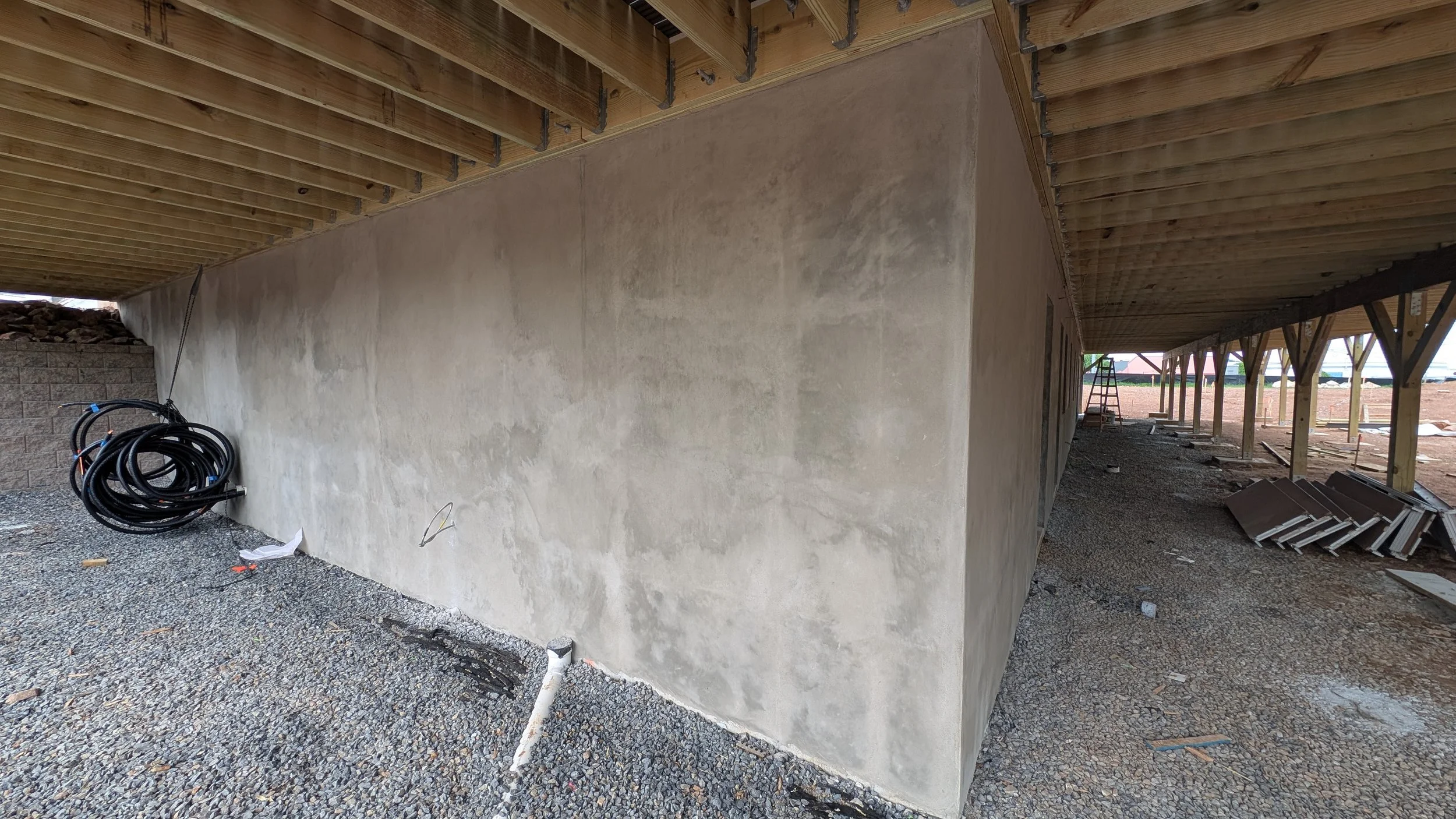 Construction site under an elevated structure with a concrete wall, gravel ground, and construction materials including coiled hoses and a ladder.