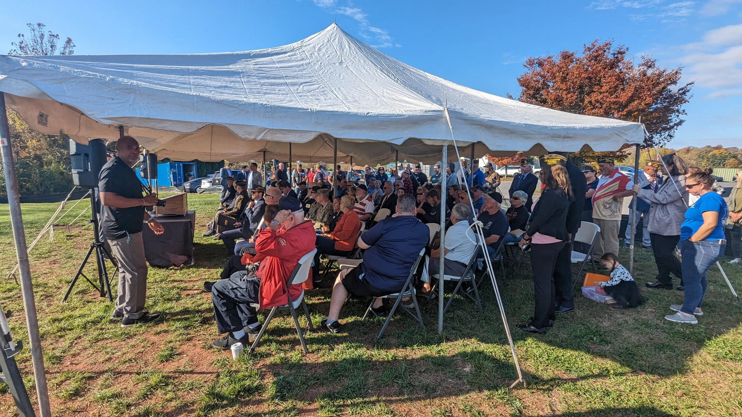 A large outdoor gathering under a white canopy tent, with attendees seated and standing, listening to a speaker at a podium. The event appears to be a formal or commemorative gathering on a sunny day with autumn foliage in the background.