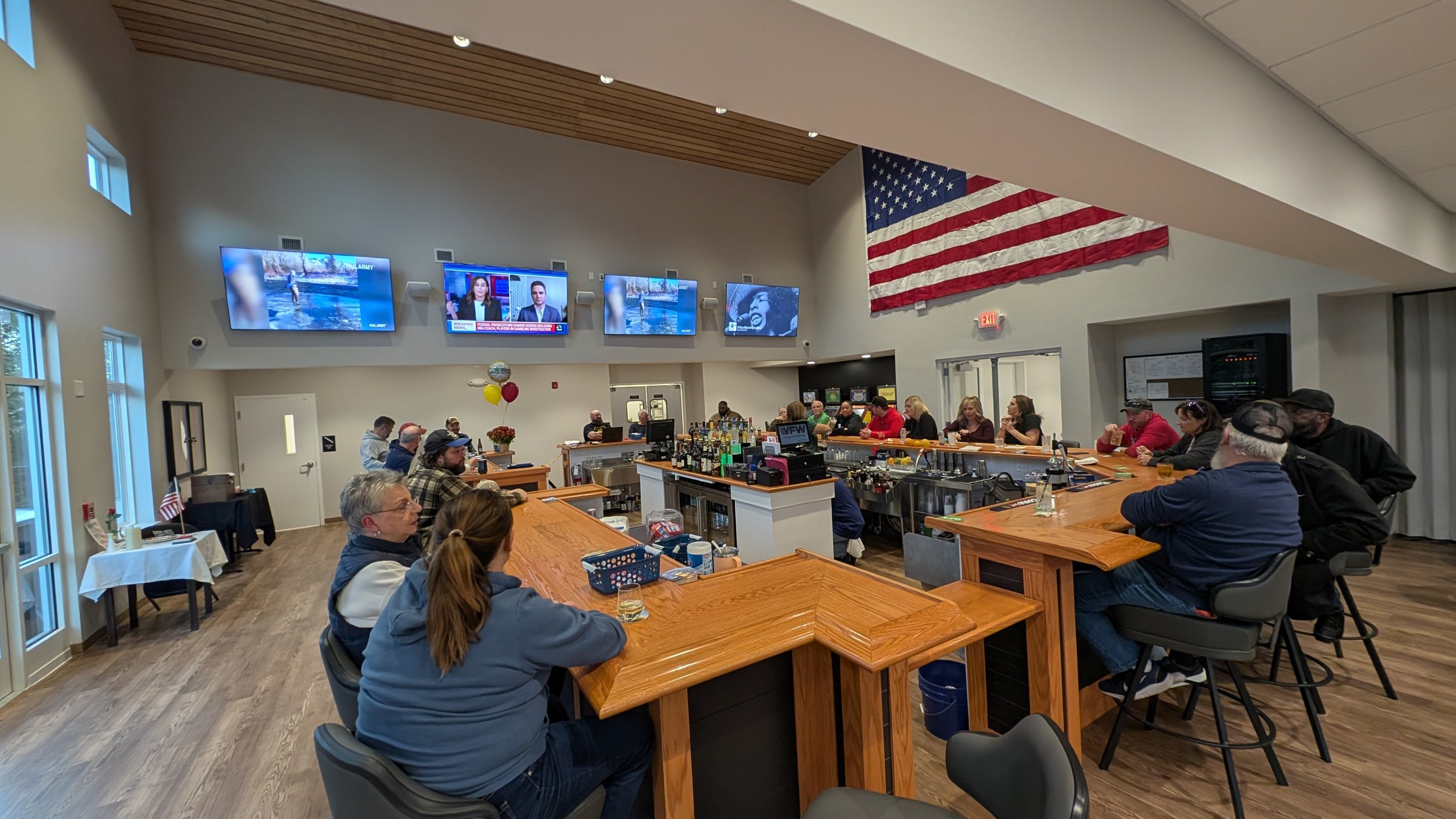 A group of people gathered in a bar or community space with a large wooden bar counter, TV screens displaying news and weather, and an American flag hanging on the wall. Some people are sitting at the bar, others are standing and socializing, and the