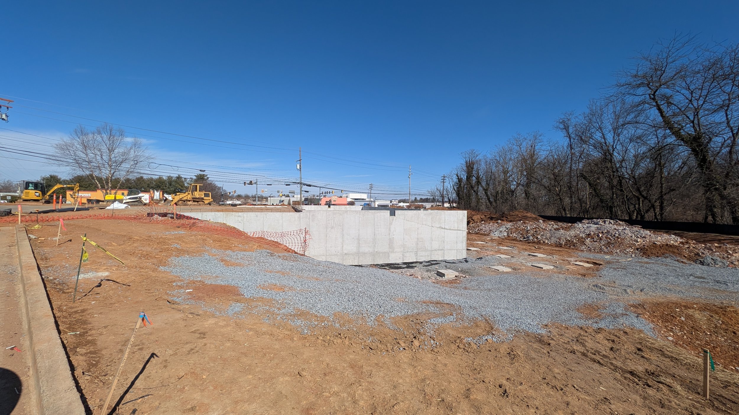 Construction site with a concrete wall, orange safety fencing, gravel, and construction equipment on a clear sunny day.