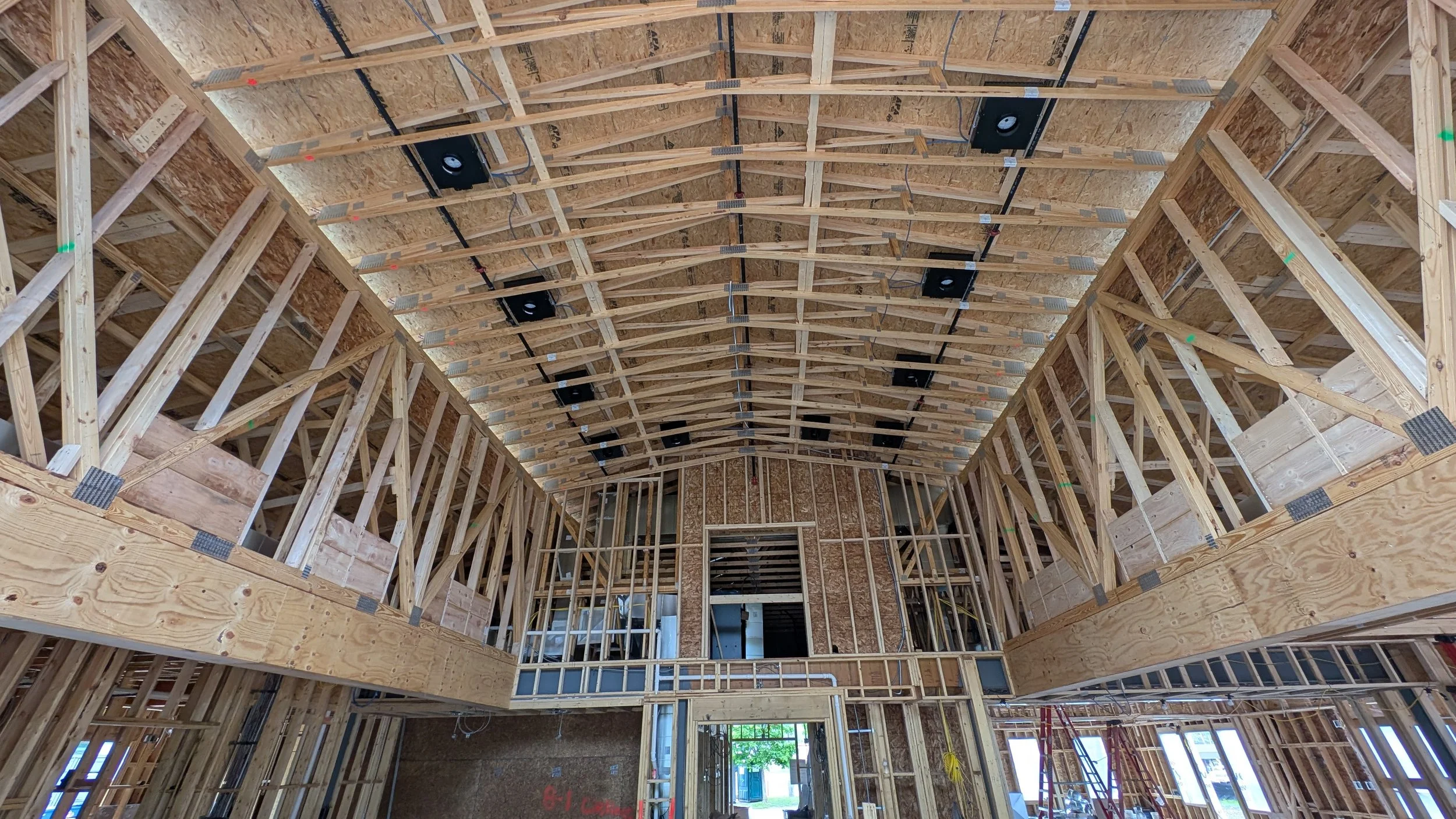 Interior view of a building under construction with wooden framing, exposed roof trusses, and some windows and doorways installed.