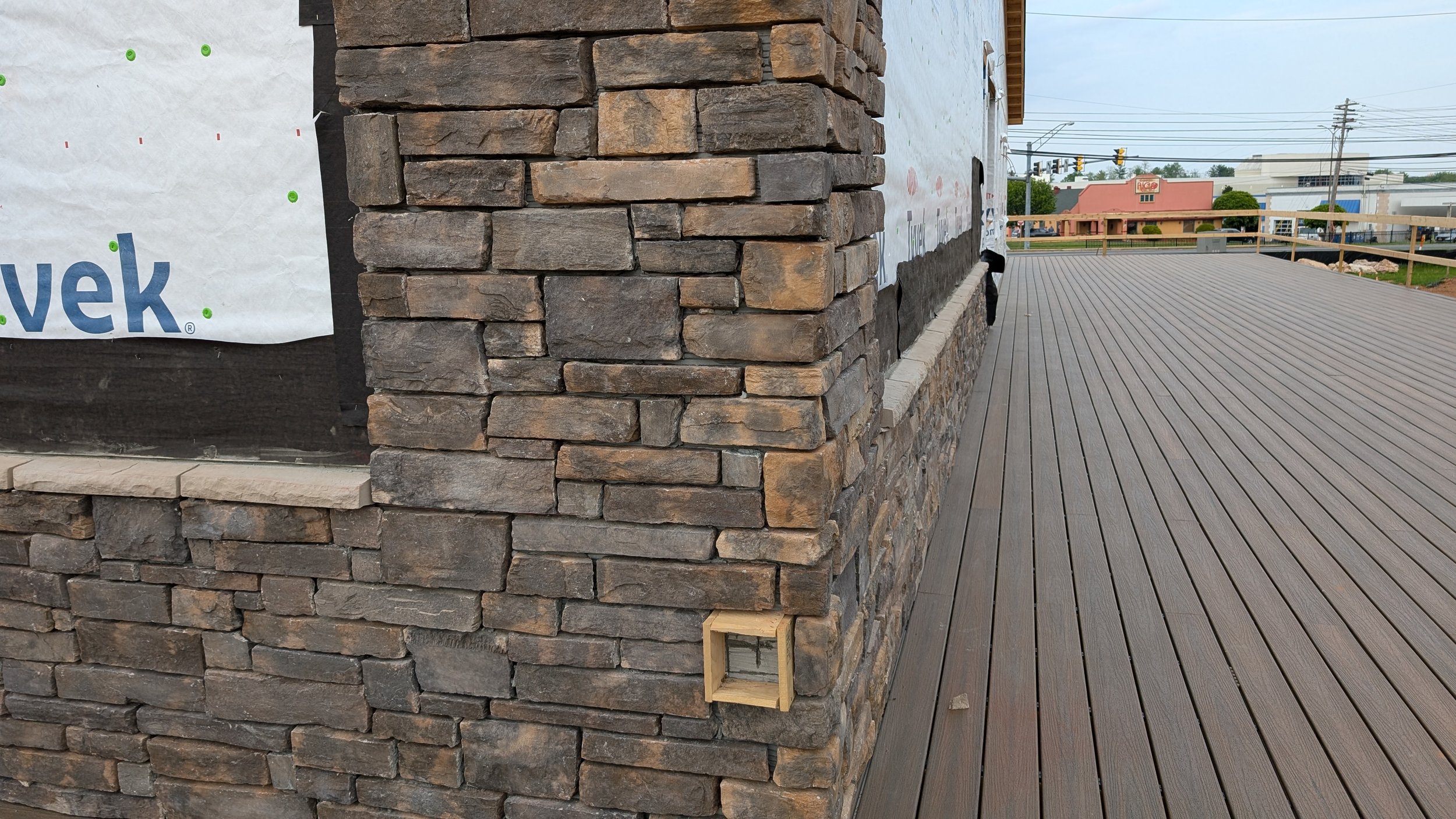 Corner of a building with a stone brick facade and a partial view of a wooden deck, with a small wooden frame attached to the brick wall near the bottom right.