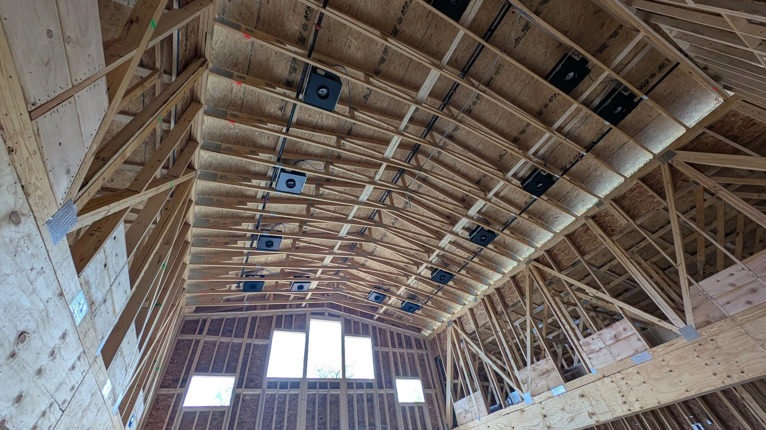 Interior view of a building under construction showing wooden framing, electrical wires, and speaker systems installed in the ceiling.