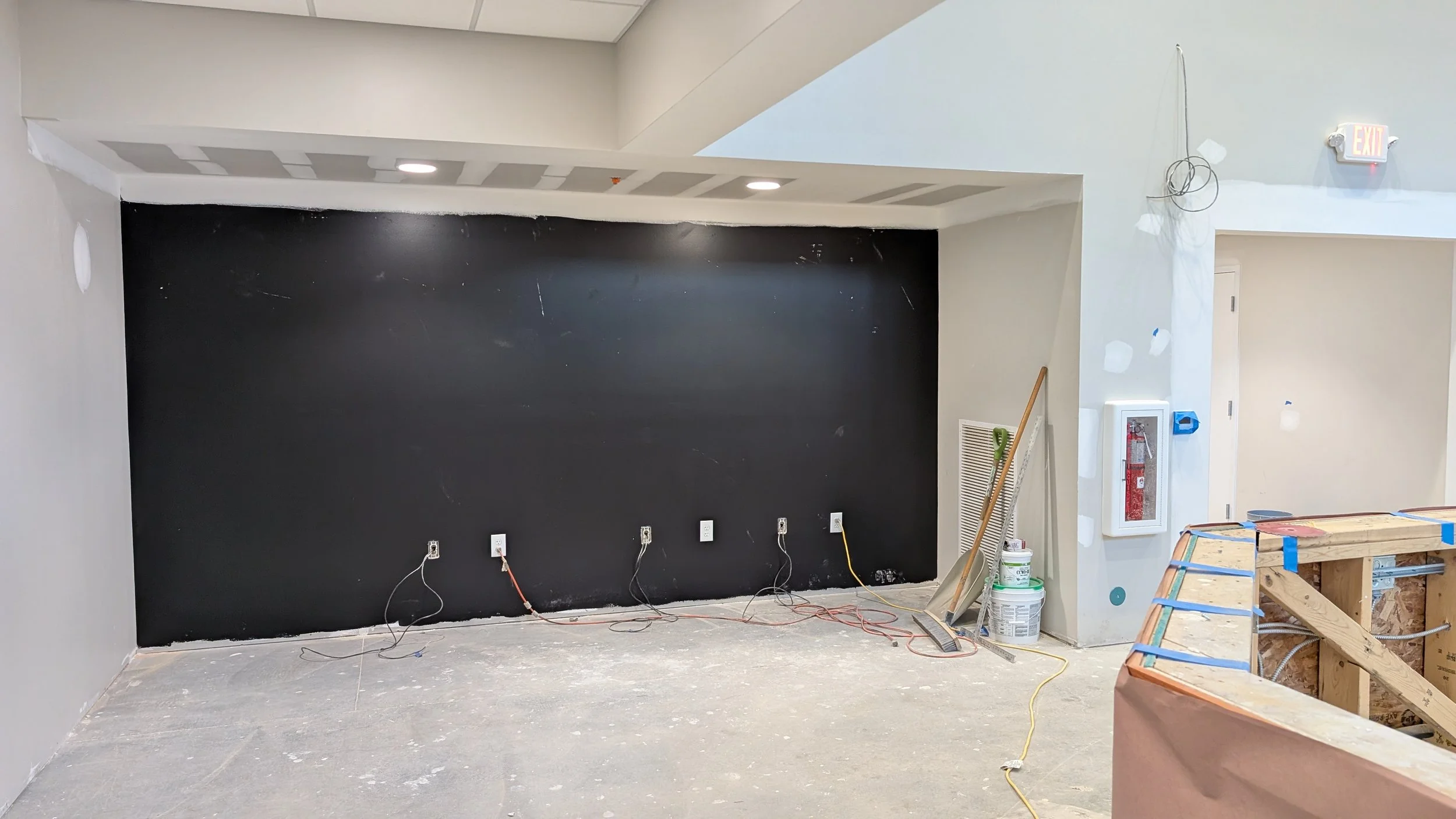 Interior view of a room under construction with a black accent wall, exposed electrical outlets and wiring, construction tools and materials on the floor, and a partially built wooden structure in the foreground.
