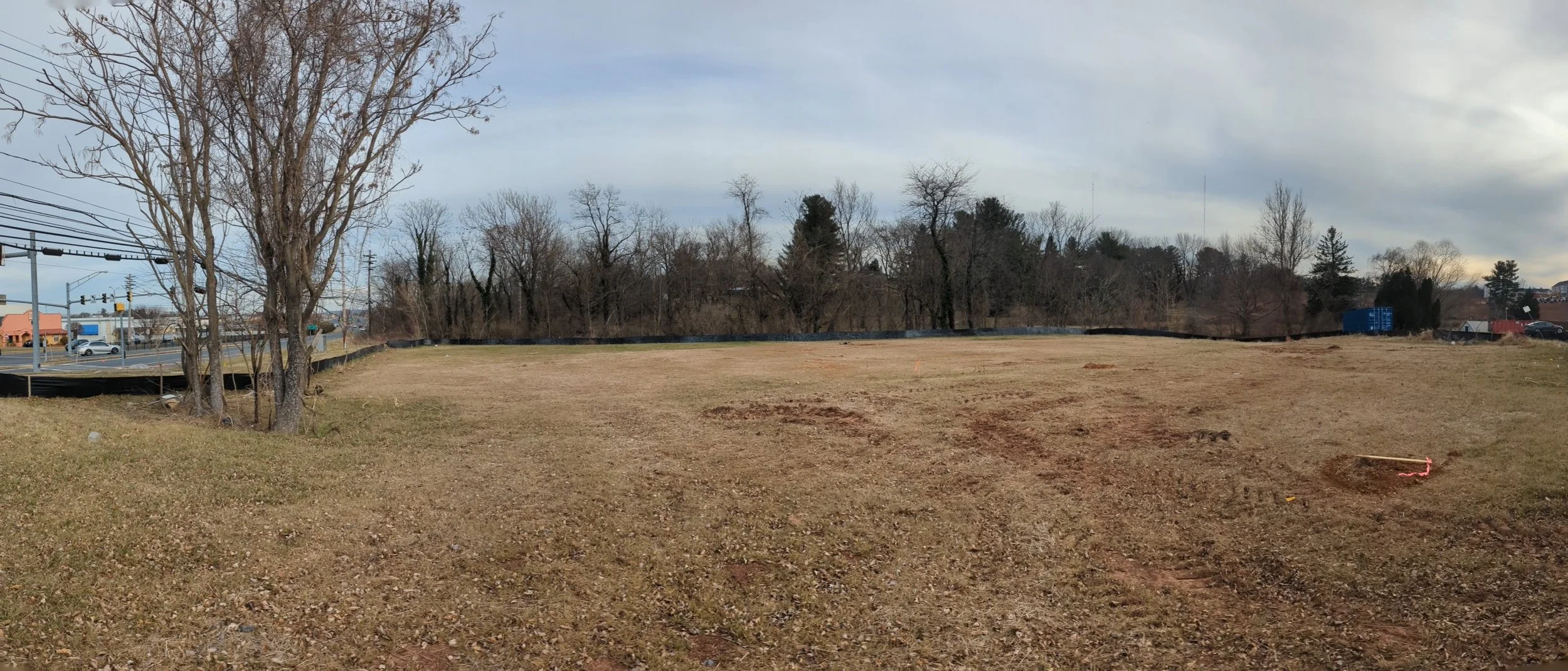 Empty lot with bare grass and dirt, trees in the background, overcast sky, some construction marks on the ground.