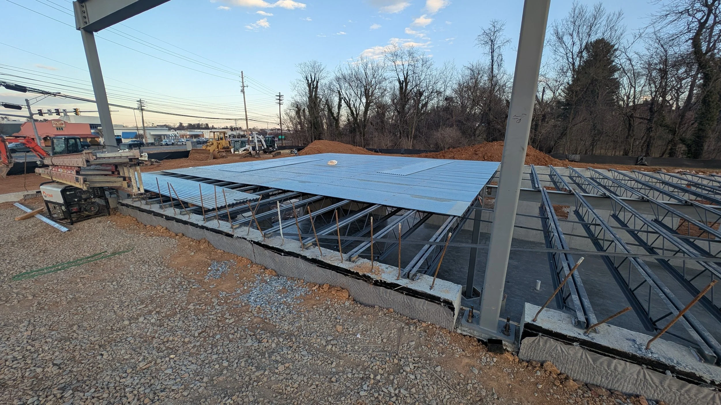 Construction site with steel framing and corrugated metal roofing panels being installed. Equipment and dirt mounds are visible in the background.