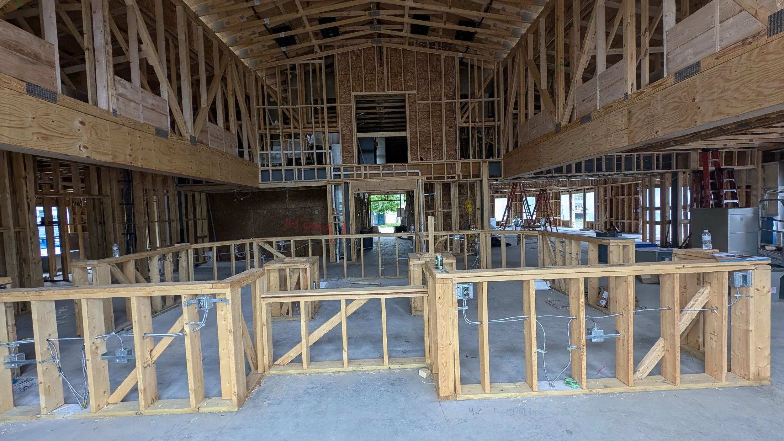 Interior view of a building under construction with wooden framing, exposed electrical wiring, scaffolding, and building materials.