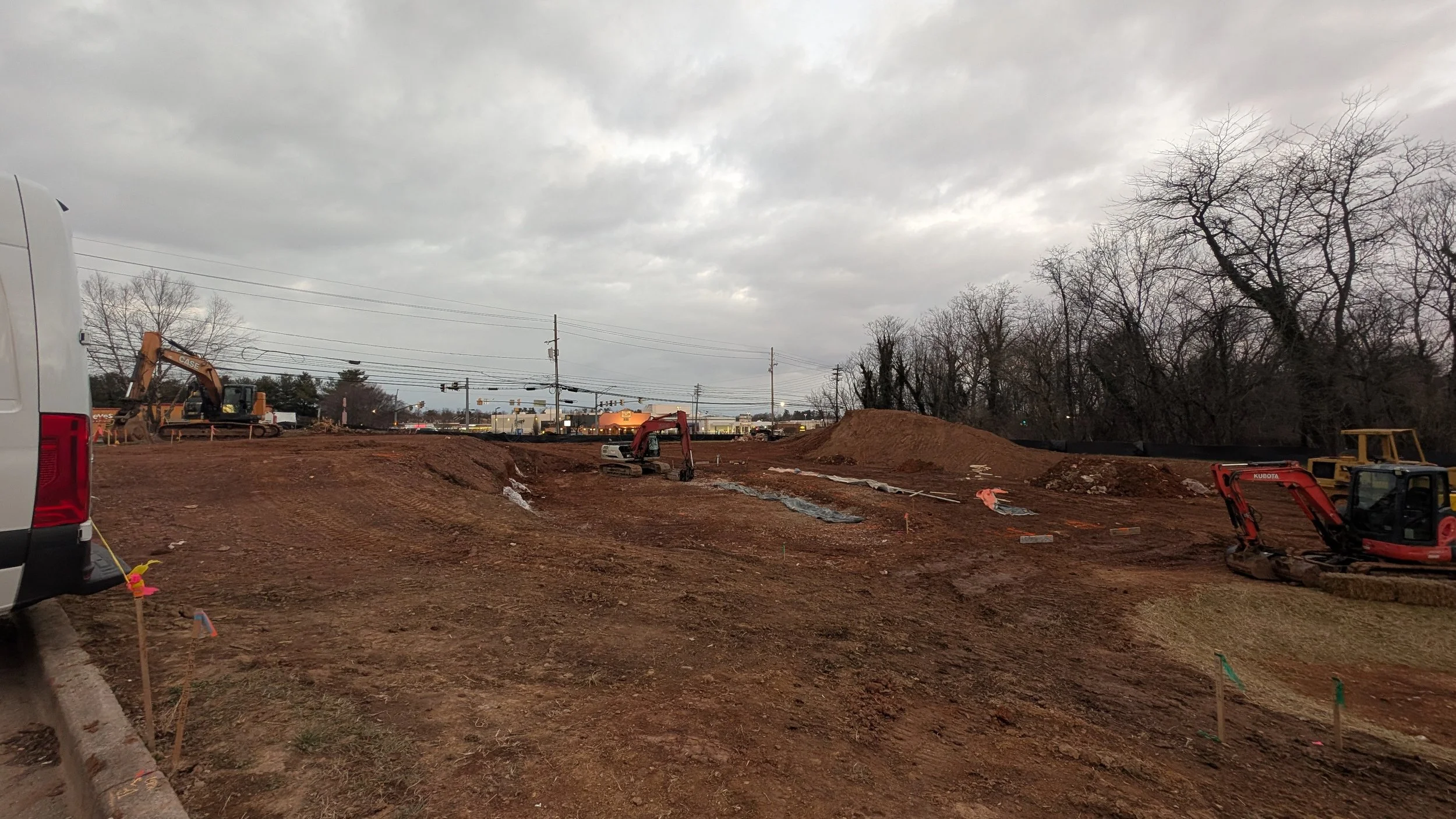 Construction site with excavators working on the dirt ground, outline of nearby trees without leaves, and vehicles and buildings in the background under an overcast sky.