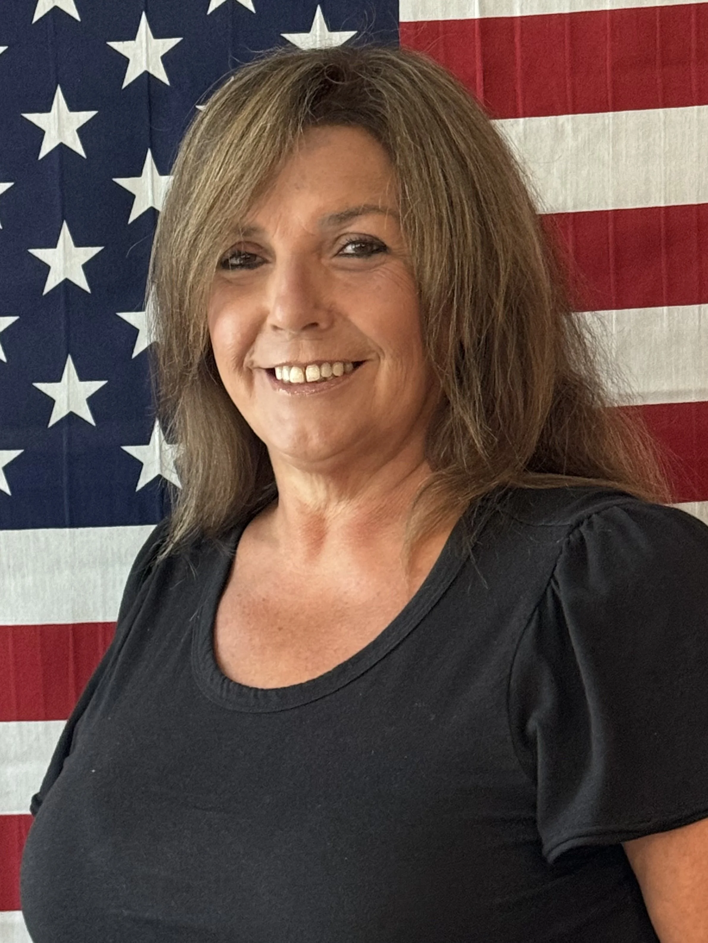 Smiling woman with shoulder-length brown hair and a black top, standing in front of an American flag.