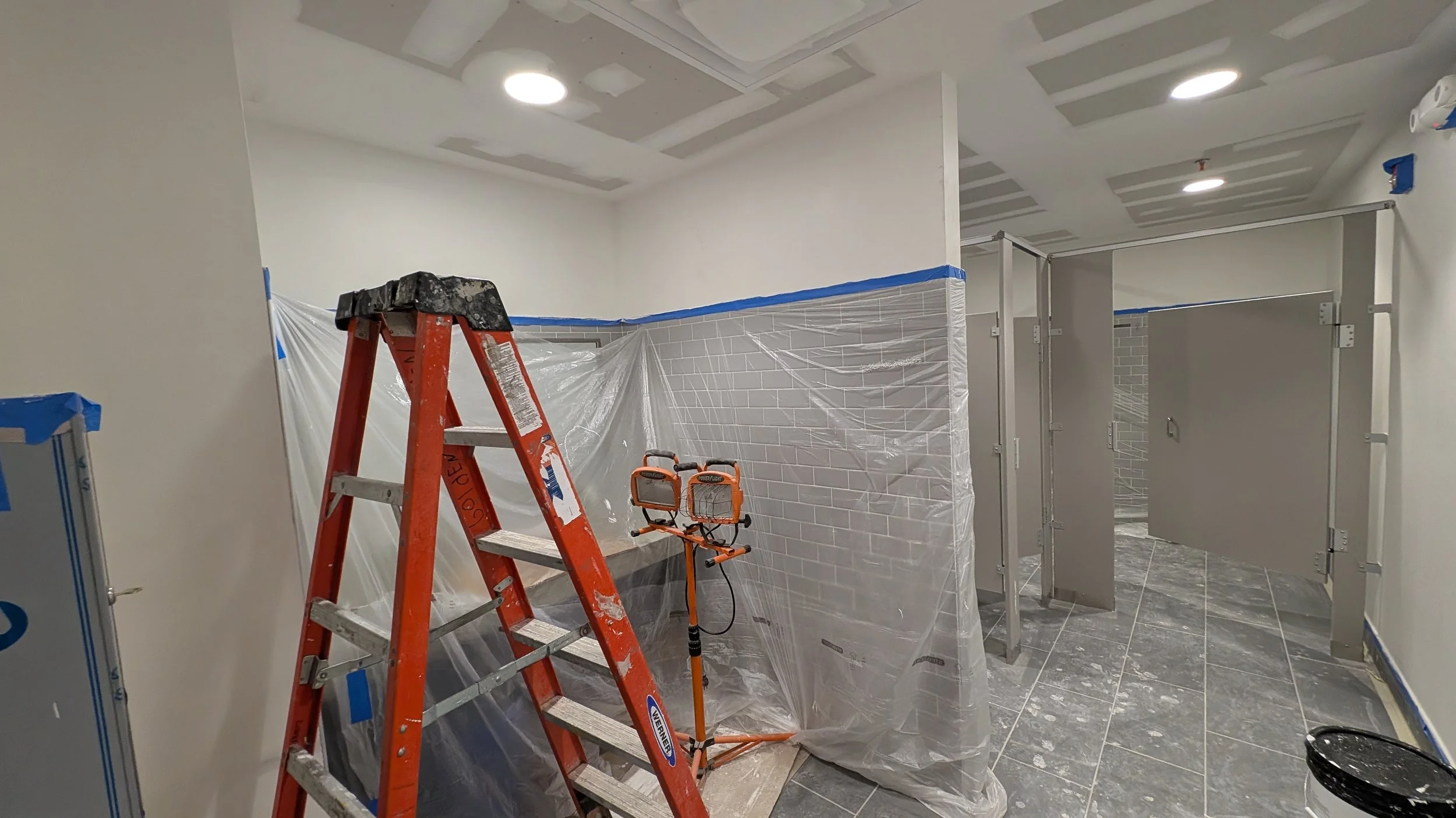 Construction site of a bathroom with drywall installation, a ladder, protective plastic sheets, and construction equipment.