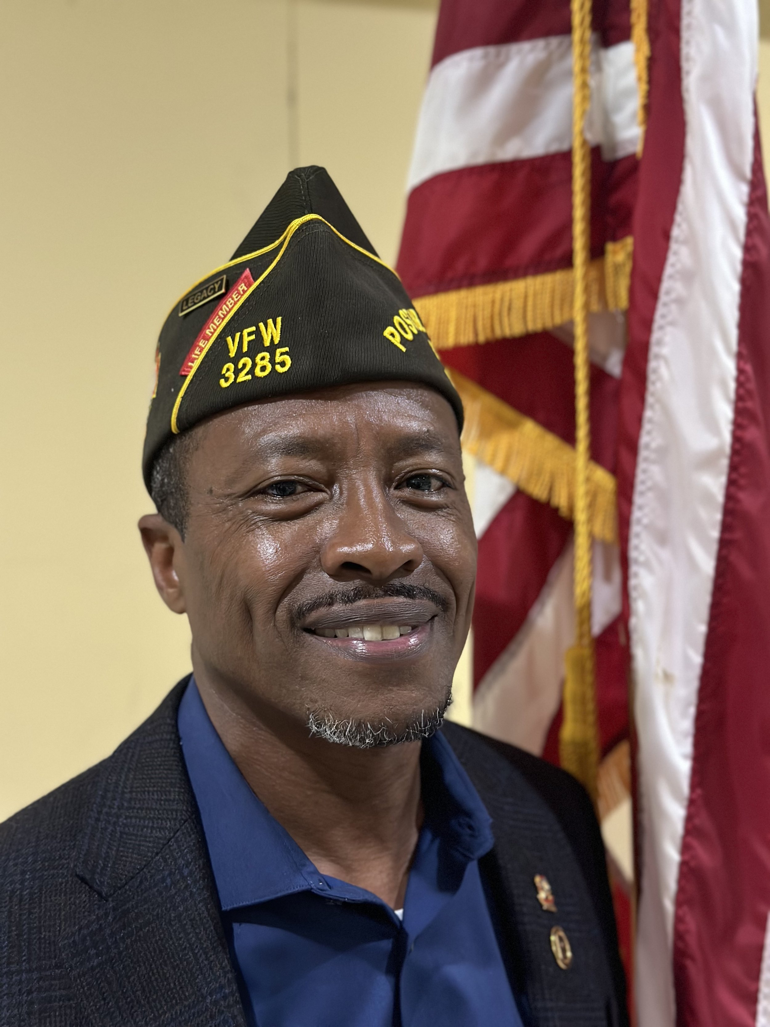 A man wearing a black Veterans of Foreign Wars (VFW) cap with yellow lettering and a gray beard, smiling in front of an American flag.