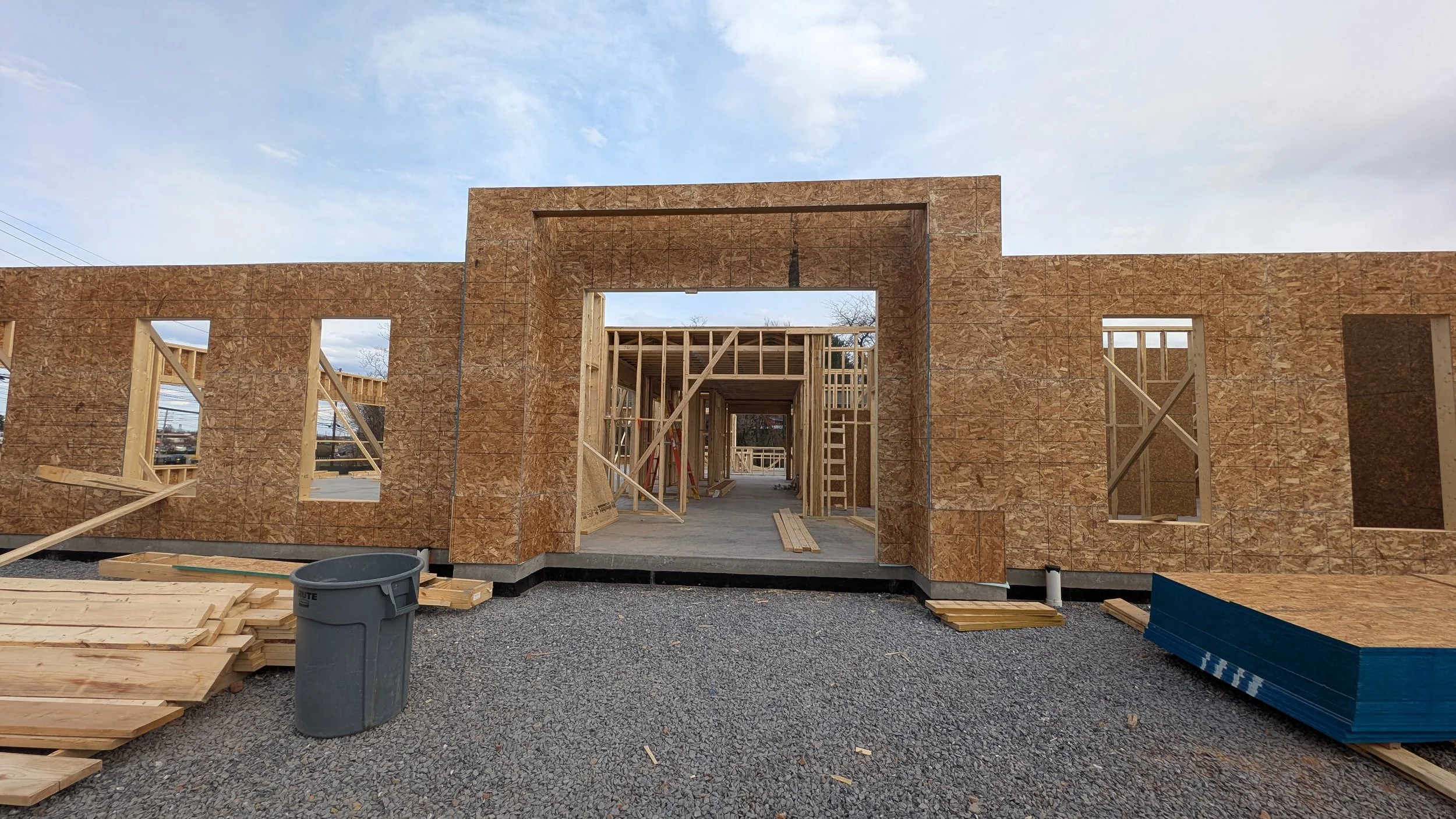 Construction site of a house with wooden framework and plywood walls, open door and window openings, gravel ground, and construction materials nearby.