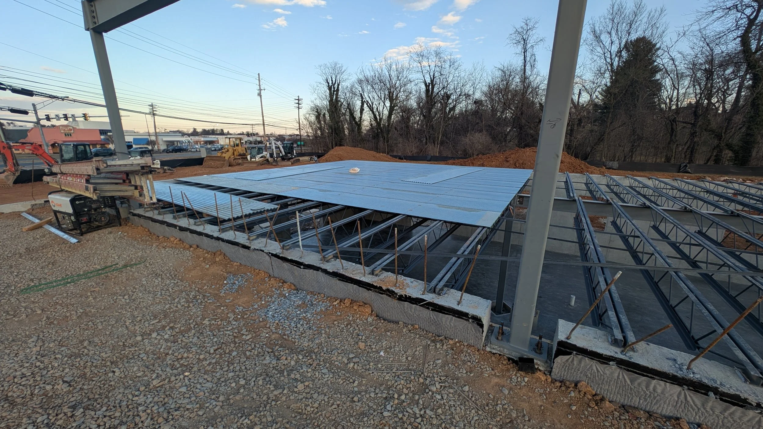 Construction site with steel framework and partially installed metal roofing panels, dirt ground, machinery in background, and trees beyond.