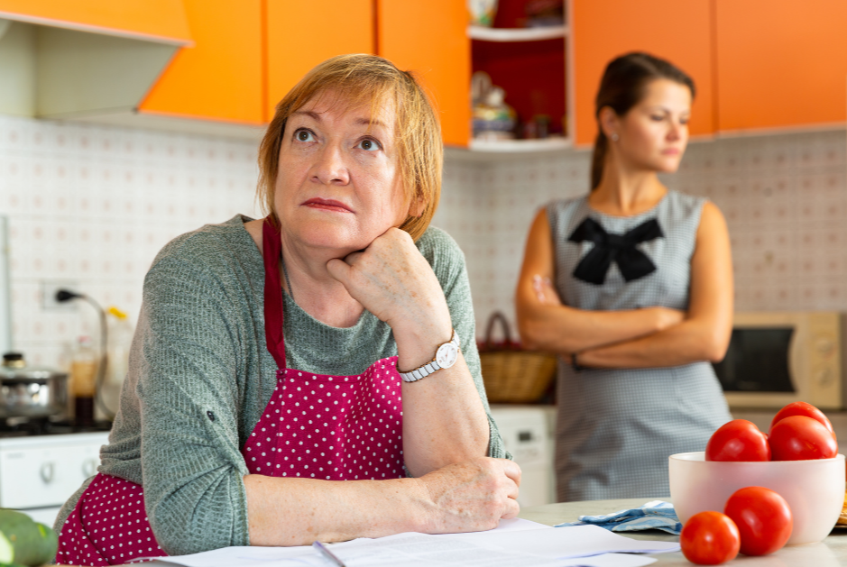 Two women in a kitchen, one older woman wearing a pink polka dot apron sitting at the counter with a thoughtful expression, and a younger woman in the background with crossed arms, looking away.