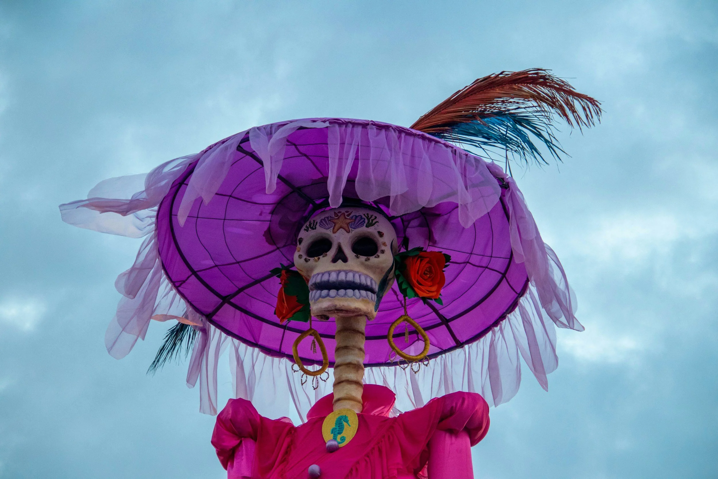 Colorful Day of the Dead skeleton with a large pink hat adorned with roses and feathers.