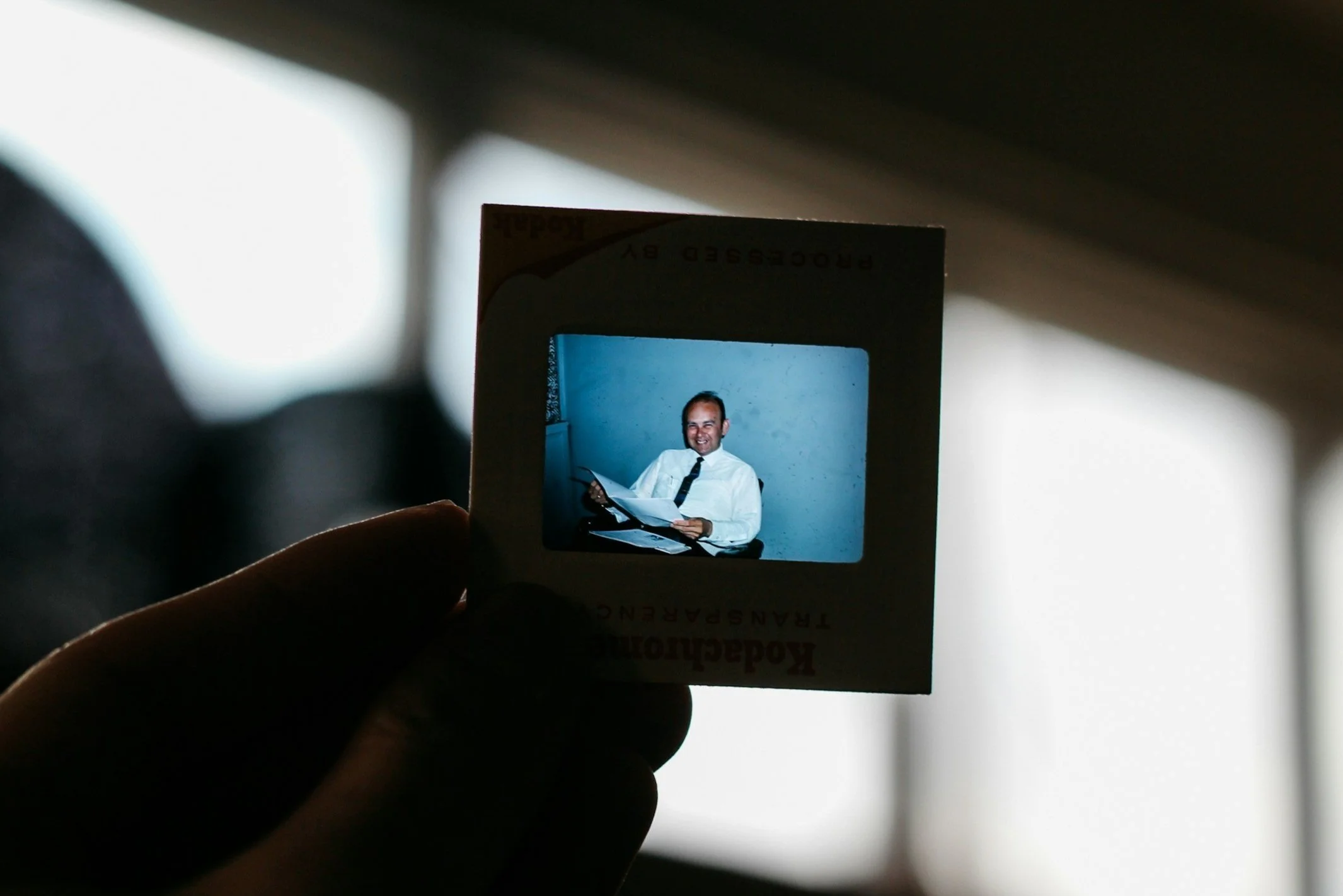 A person holds a slide of a photograph showing a man in a white shirt and black tie smiling while sitting at a desk with papers.