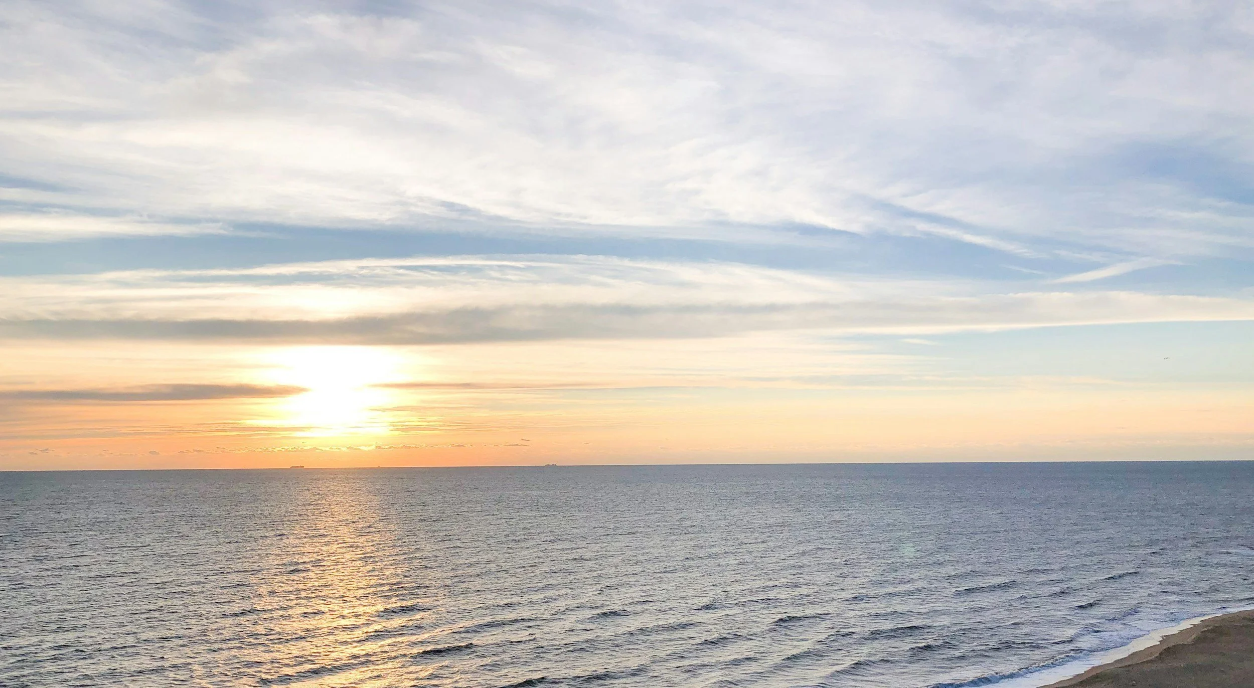 Sunset over a calm ocean with clear skies and gentle waves near a sandy beach.